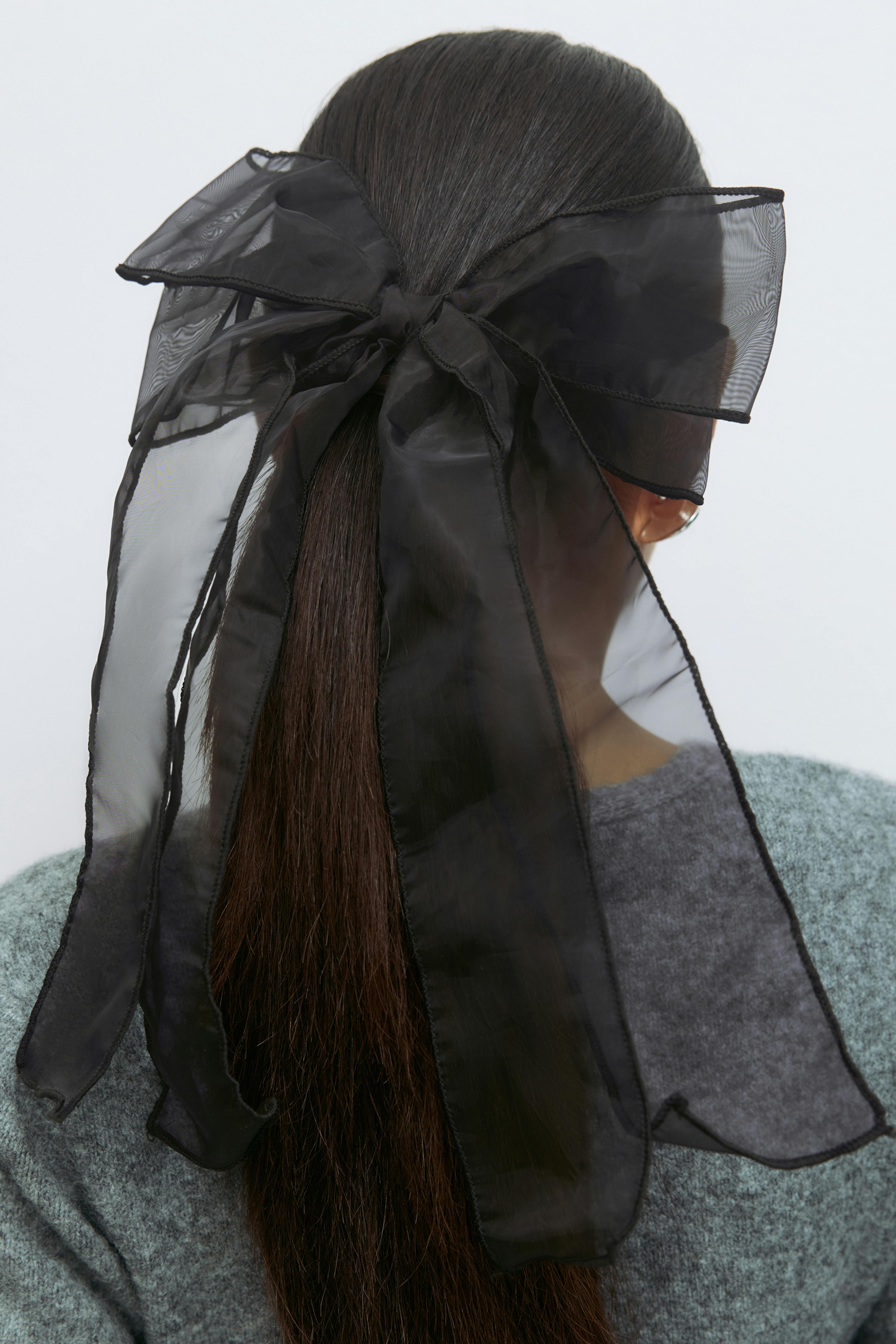 Sheer black organza hair bow with long trailing ends tied over a low ponytail; grey textured knit sweater.