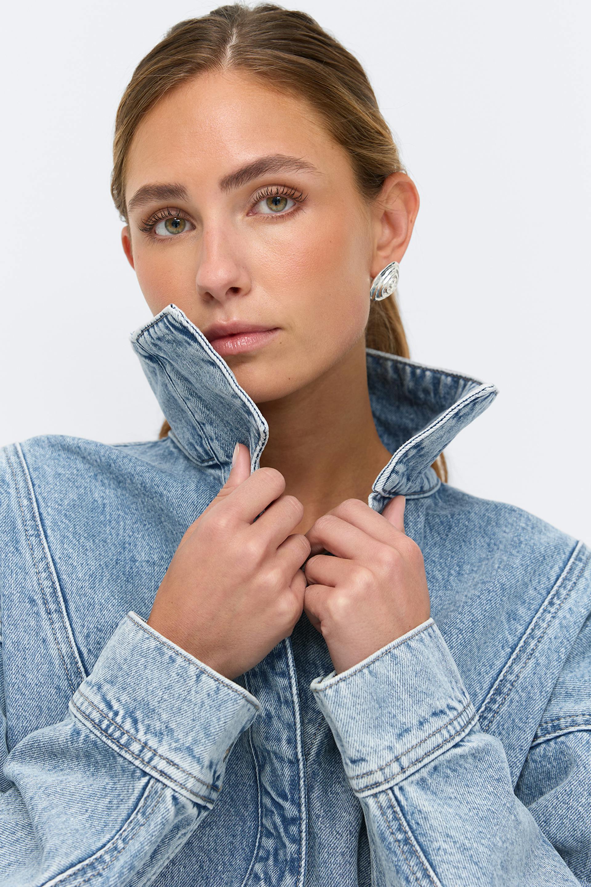 Kevyesti pesty farkkutakki, jossa on ylöskäännetty kaulus ja kontrastiompeleet; hopeiset veistokselliset korvakorut.