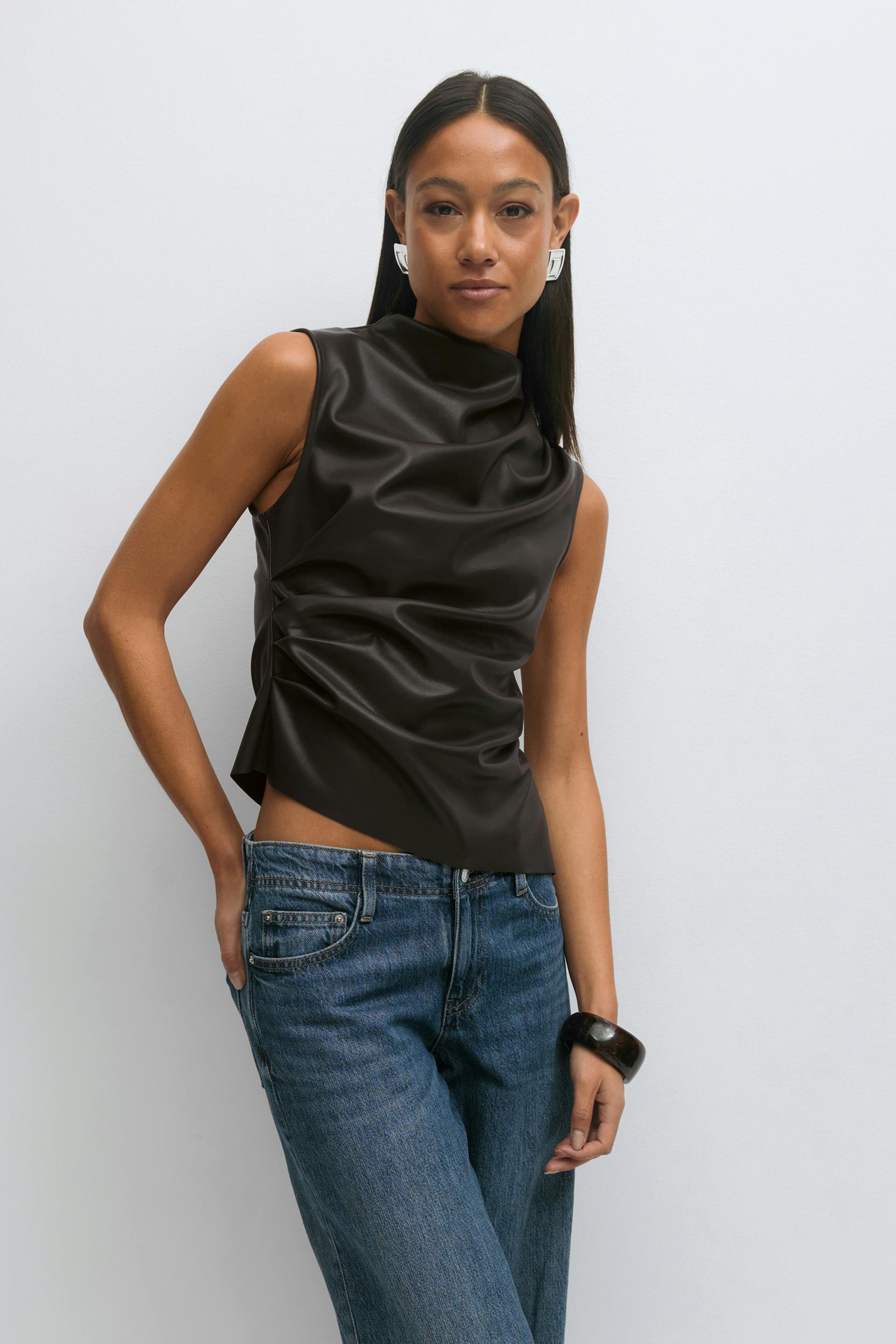 Sleeveless dark brown faux‑leather draped cowl‑neck top with an asymmetrical hem, paired with medium‑wash straight‑leg jeans; styled with white geometric earrings and a black chunky bangle.