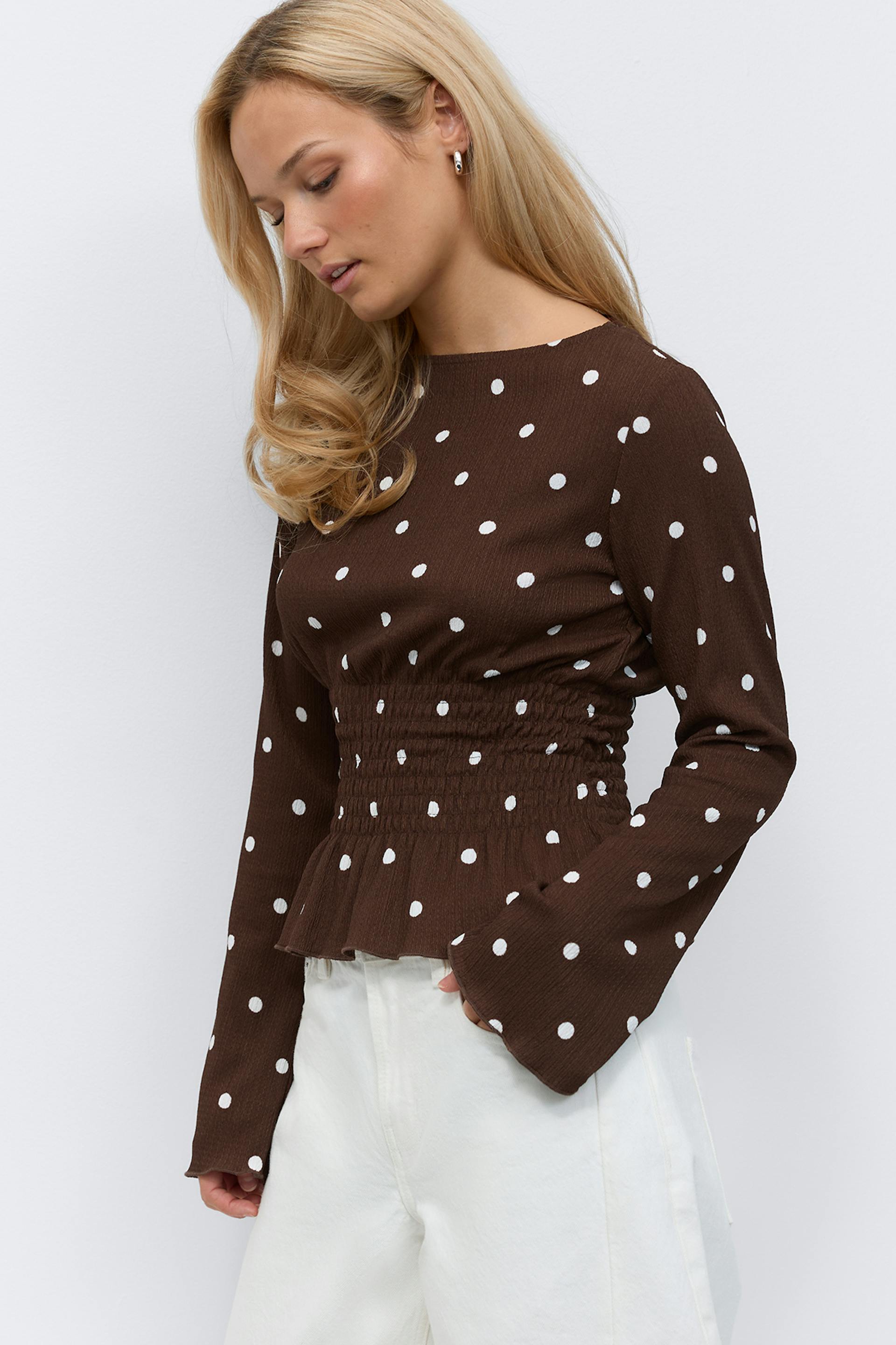 Ruskea pitkähihainen pusero, jossa valkoisia pilkkuja, smokattu vyötärö ja hienovarainen peplum-helma, kellohihansuut; yhdistettynä korkeavyötäröisiin valkoisiin leveälahkeisiin housuihin.