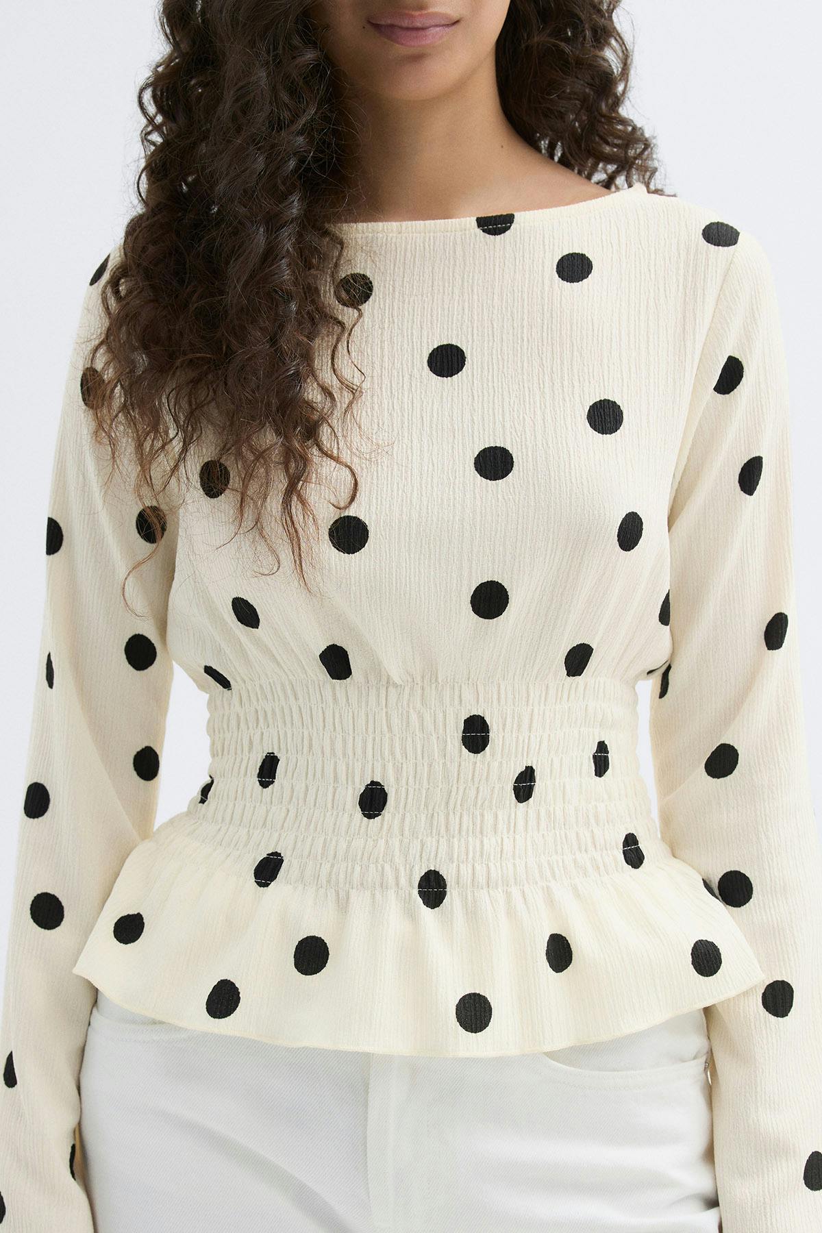Cremefarbene, strukturierte Langarmbluse mit schwarzen Tupfen, Bootsausschnitt, gesmokter Taille und Schößchensaum, kombiniert mit einer weißen Hose.