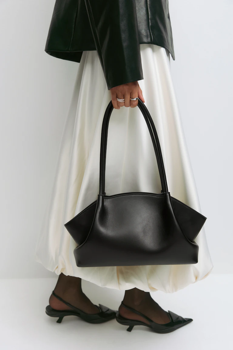 Svart skinnjacka, kjol i elfenbensfärgad satin med ballongfåll, skira svarta strumpbyxor och svarta spetsiga slingback-kitten heels; bär en strukturerad svart skinnhandväska och silverringar.