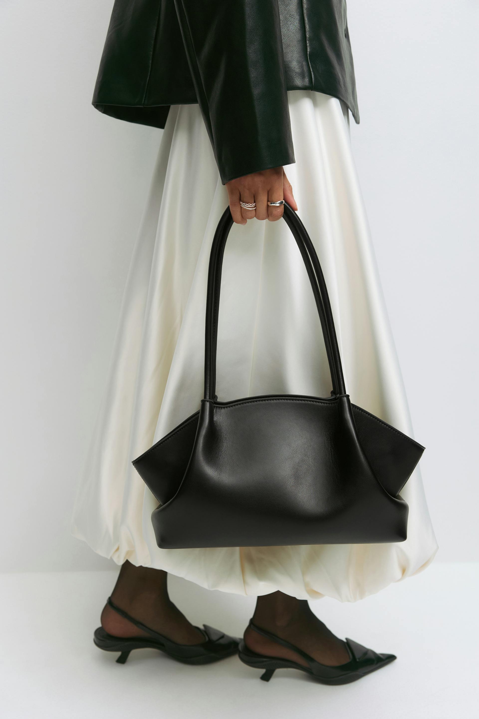 Black leather jacket, ivory satin full skirt with balloon hem, sheer black tights and black pointed slingback kitten heels; carrying a structured black leather handbag and wearing silver rings.