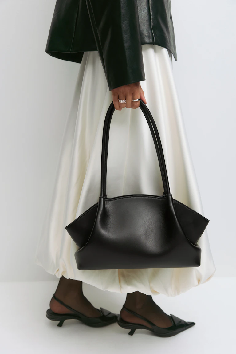 Svart skinnjacka, kjol i elfenbensfärgad satin med ballongfåll, skira svarta strumpbyxor och svarta spetsiga slingback-kitten heels; bär en strukturerad svart skinnhandväska och silverringar.