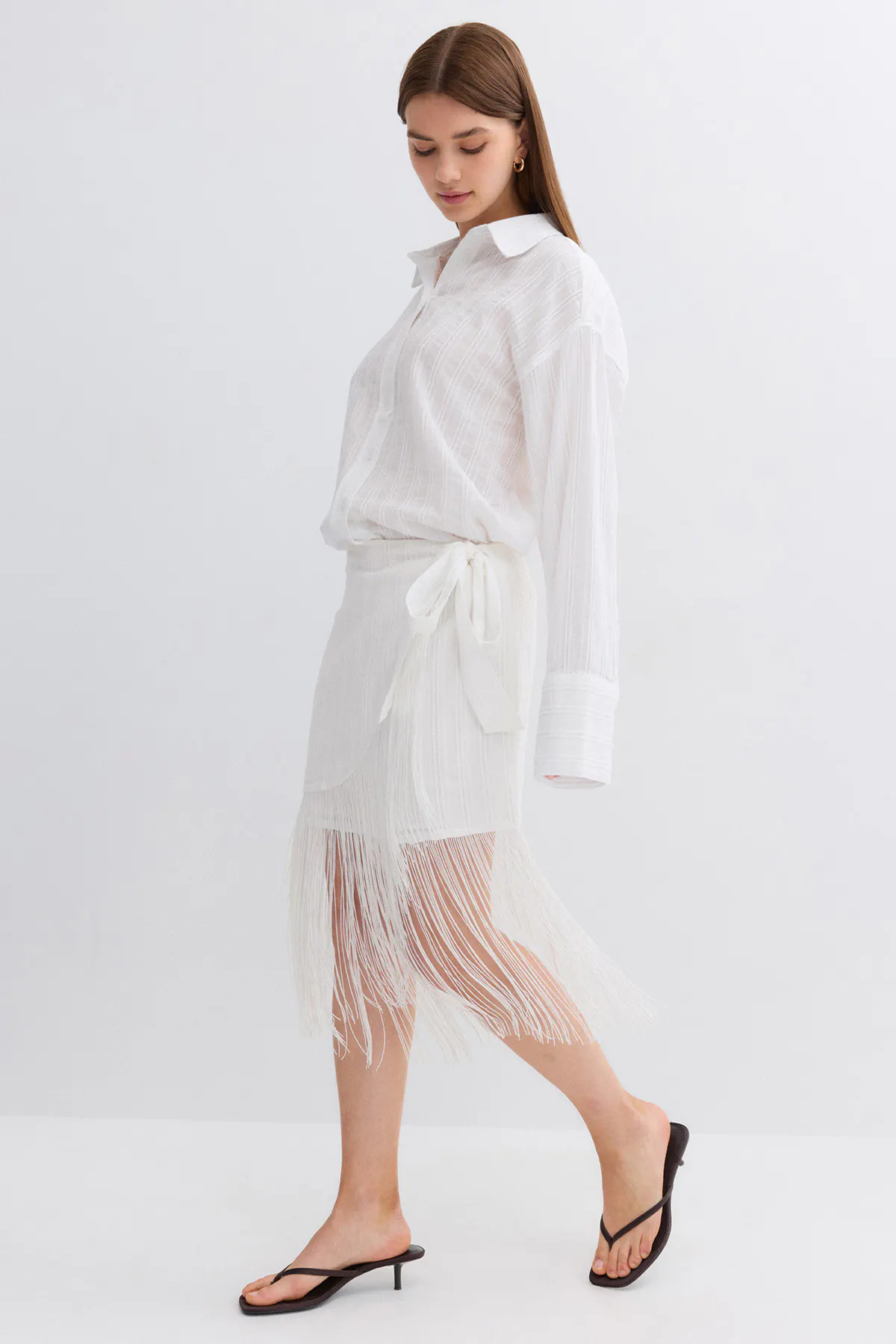 Valkoinen pitkähihainen, napitettava, pitkähihainen pusero kevyestä kuvioidusta kankaasta, joka on sidottu vyötäröllä; yhteensopiva valkoinen kietaisuhame, jossa on sivuside ja pitkä hapsuhelmainen helma; mustat matalakorkoiset stringisandaalit.