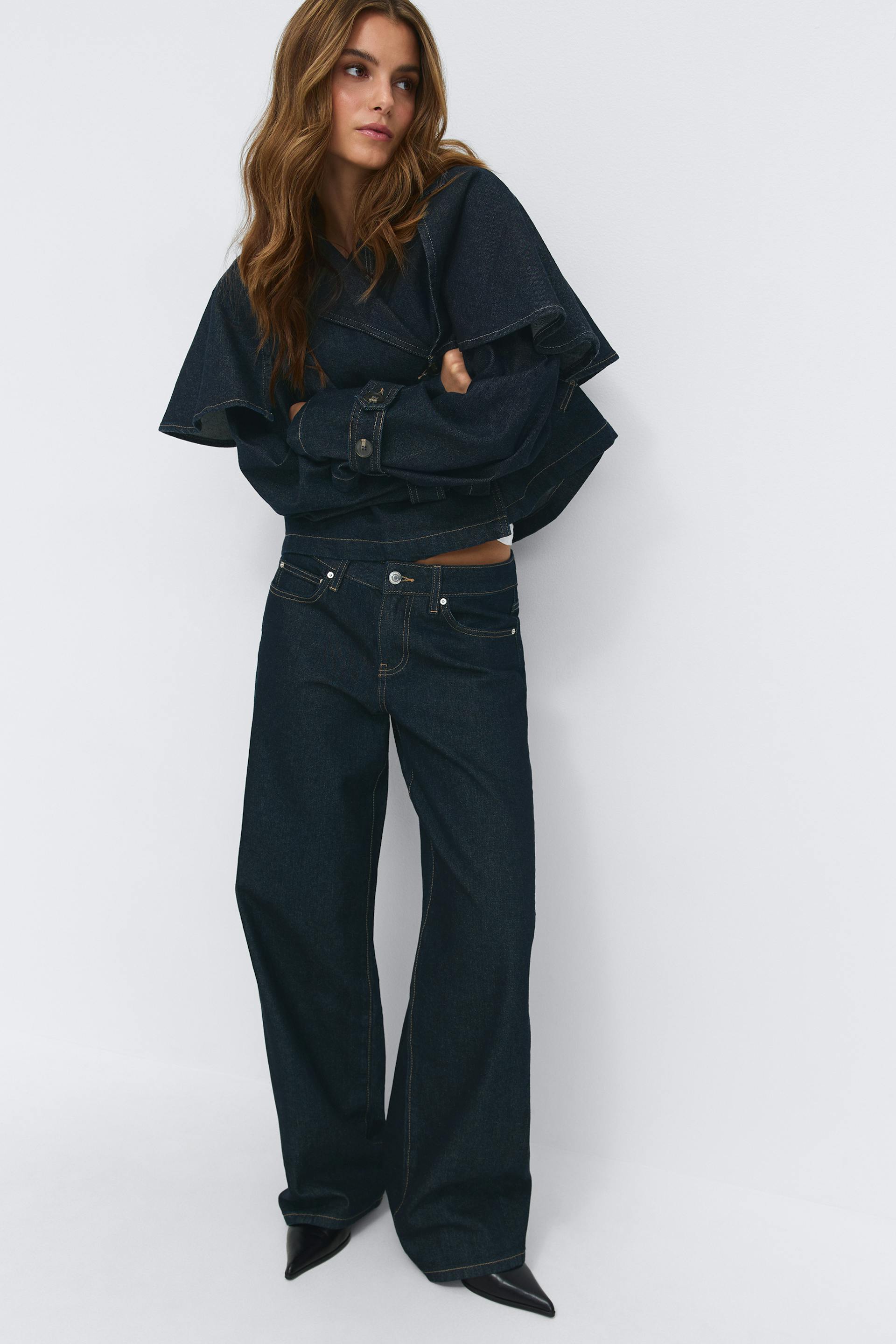 Hellrosafarbenes, langärmeliges Oversize-Button-Down-Hemd, offen über einem weißen, gerippten Tank getragen, kombiniert mit hochtaillierten, indigofarbenen Jeans mit weitem Bein, spitzen burgunderroten Absätzen und goldenen Ohrringen.