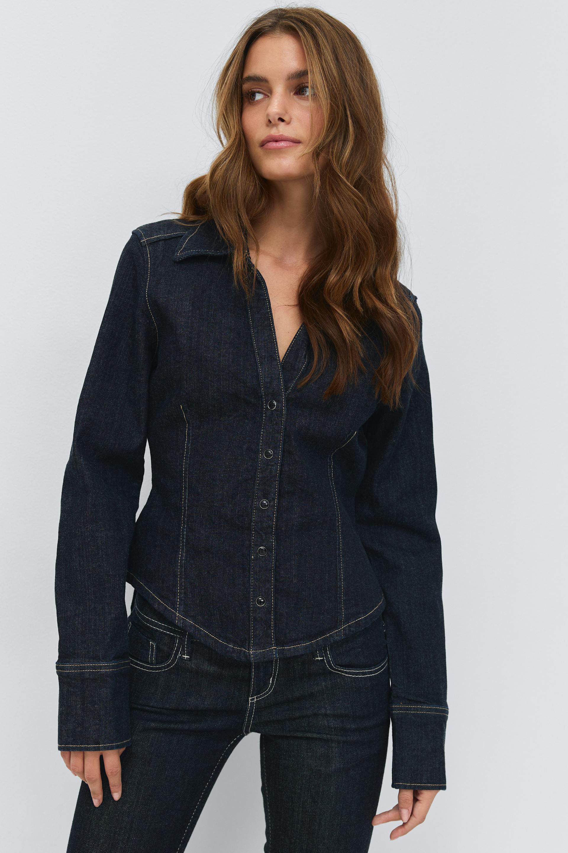 Dark indigo fitted denim button-up shirt with pointed collar, long sleeves and extended cuffs, featuring contrast topstitching; paired with matching dark indigo denim jeans with contrast stitching.
