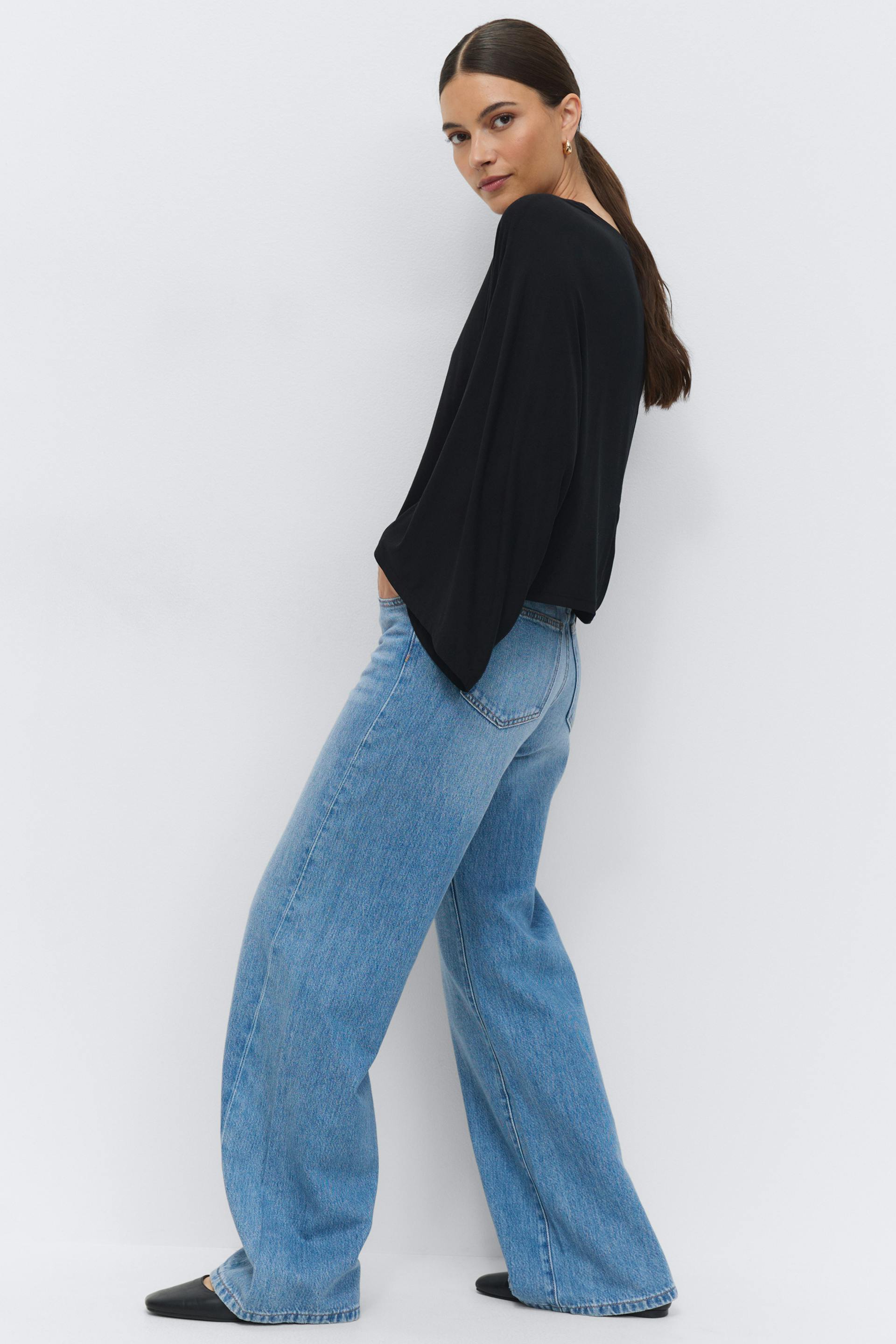 Zwarte blouse met lange mouwen en gedrapeerde wijde mouwen; lichtblauwe jeans met hoge taille en wijde pijpen; zwarte spitse platte schoenen; kleine gouden oorbellen.