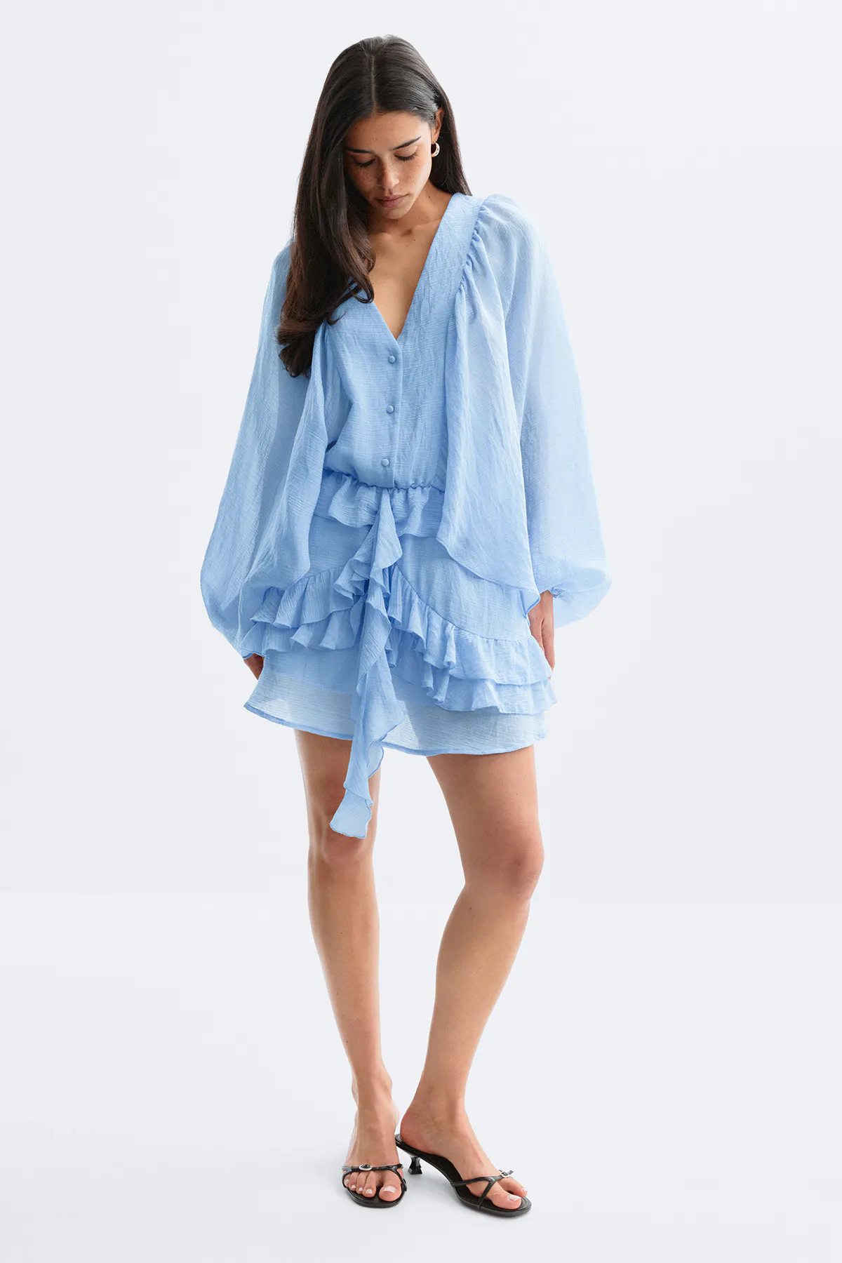 Vaaleansininen V-pääntiellä varustettu nappikiinnitteinen minimekko, jossa on pitkät hihat, solmittava vyötärö ja tiered röyhelöinen helma, yhdistettynä mustiin matalakorkoisiin sandaaleihin.