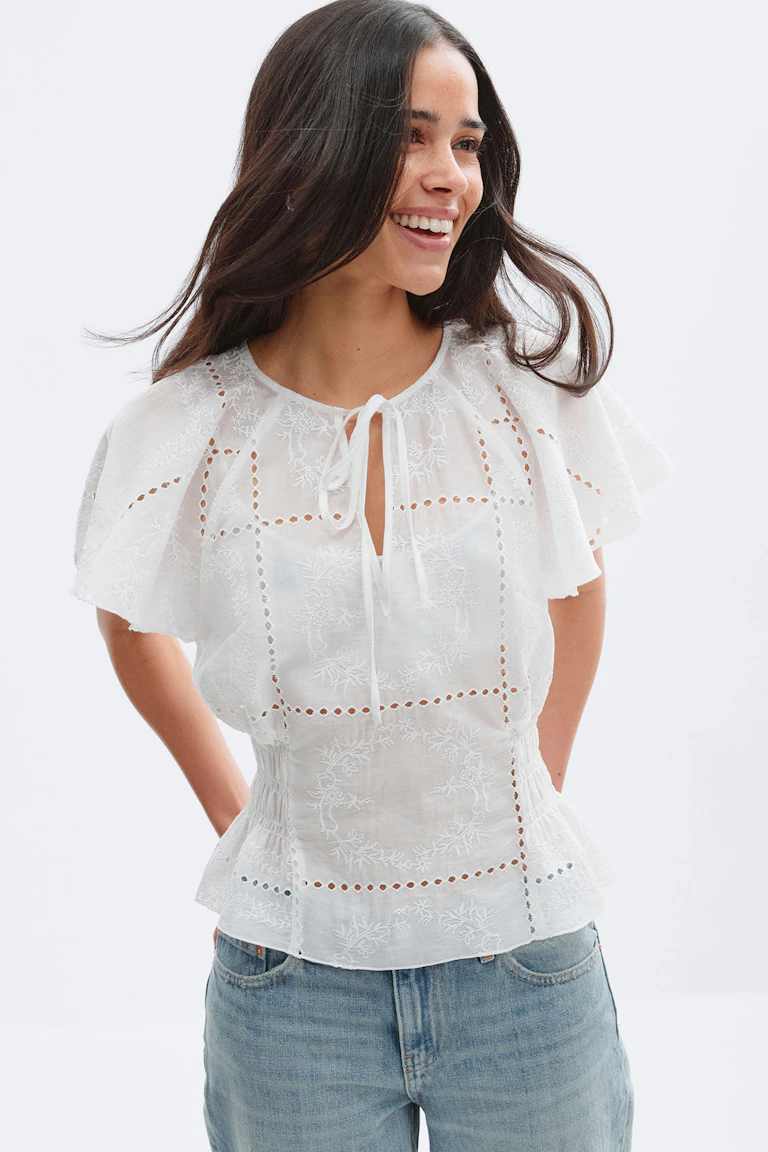 Witte blouse van geborduurd eyelet met fladdermouwen, knoopsgat aan de voorkant en gesmokte peplum in de taille, gecombineerd met een jeans van lichtgewassen denim.