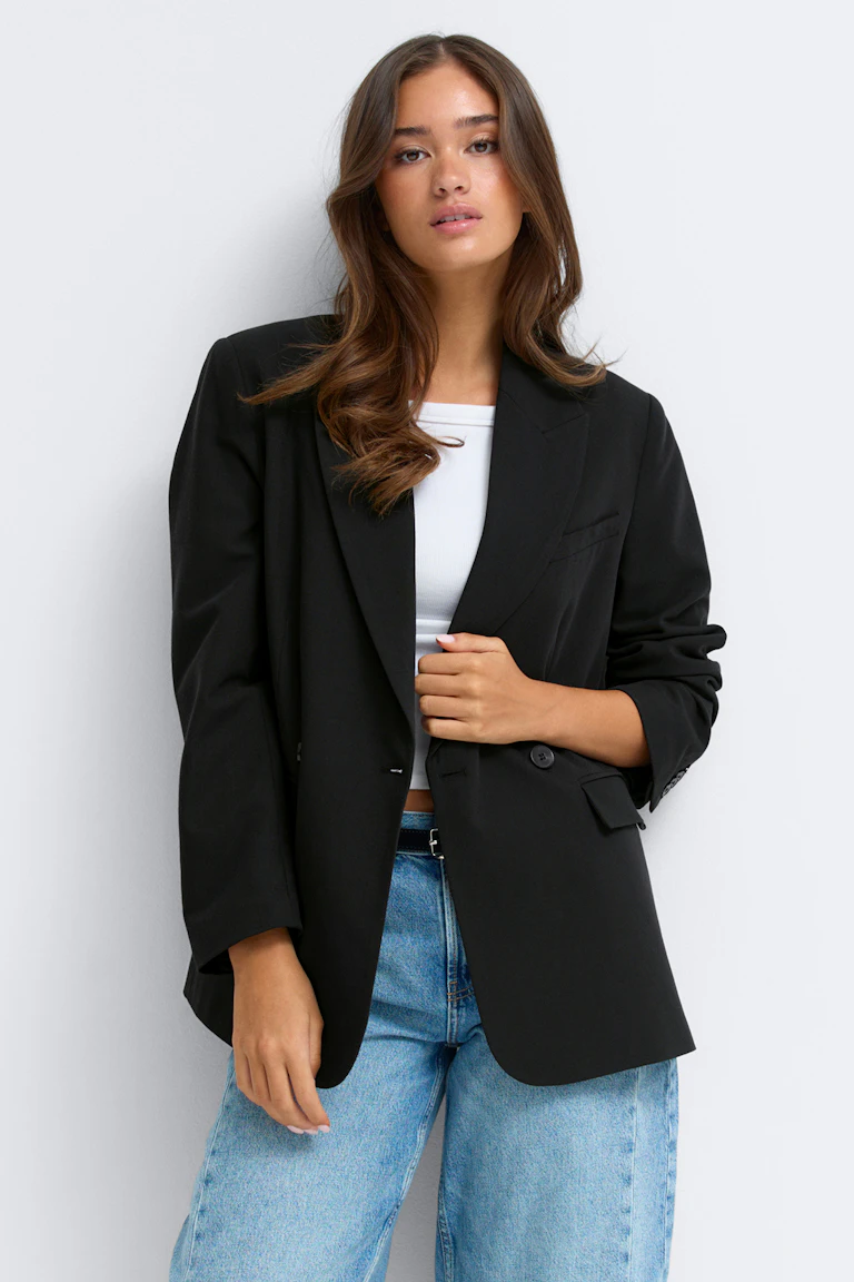 Sort oversized dobbeltradet blazer med hakkede revers og klaplommer båret over en sort top, sammen med sorte bukser med brede ben og sorte spidse sko.