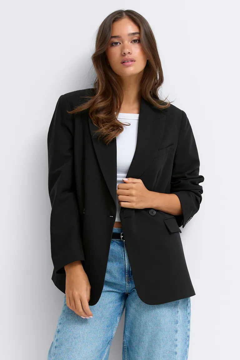 Schwarzer doppelreihiger Oversize-Blazer mit Revers und Pattentaschen, getragen über einem schwarzen Oberteil, kombiniert mit einer schwarzen Hose mit weitem Bein und schwarzen spitzen Schuhen.