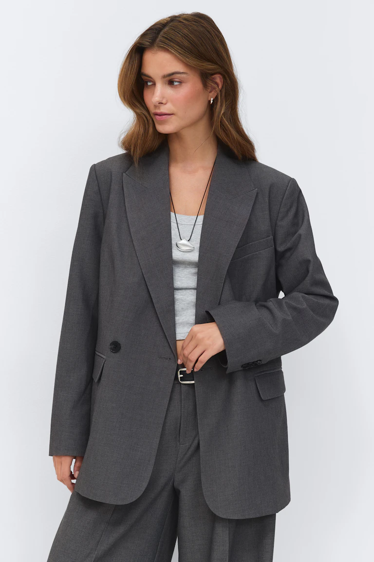 Anthrazitfarbener, einreihiger Oversize-Blazer und passende Hose mit weitem Bein, kombiniert mit einem hellgrauen, gerippten Tanktop, einem schwarzen Gürtel mit silberner Schnalle und einer silbernen Halskette mit Anhänger.