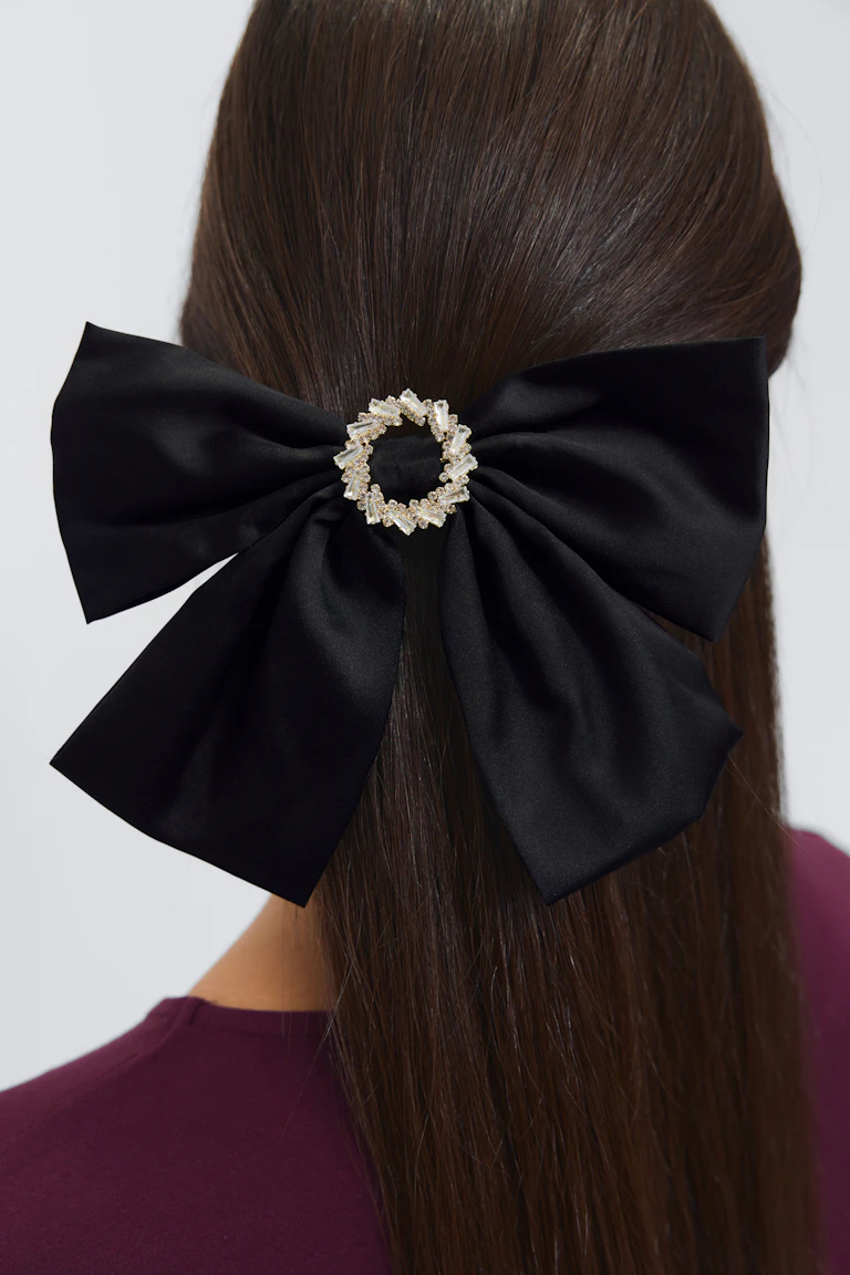 Zwart satijnen strik haaraccessoire met een ronde met kristallen versierde gouden broche in het midden, gecombineerd met een bordeauxrode crew-neck top.