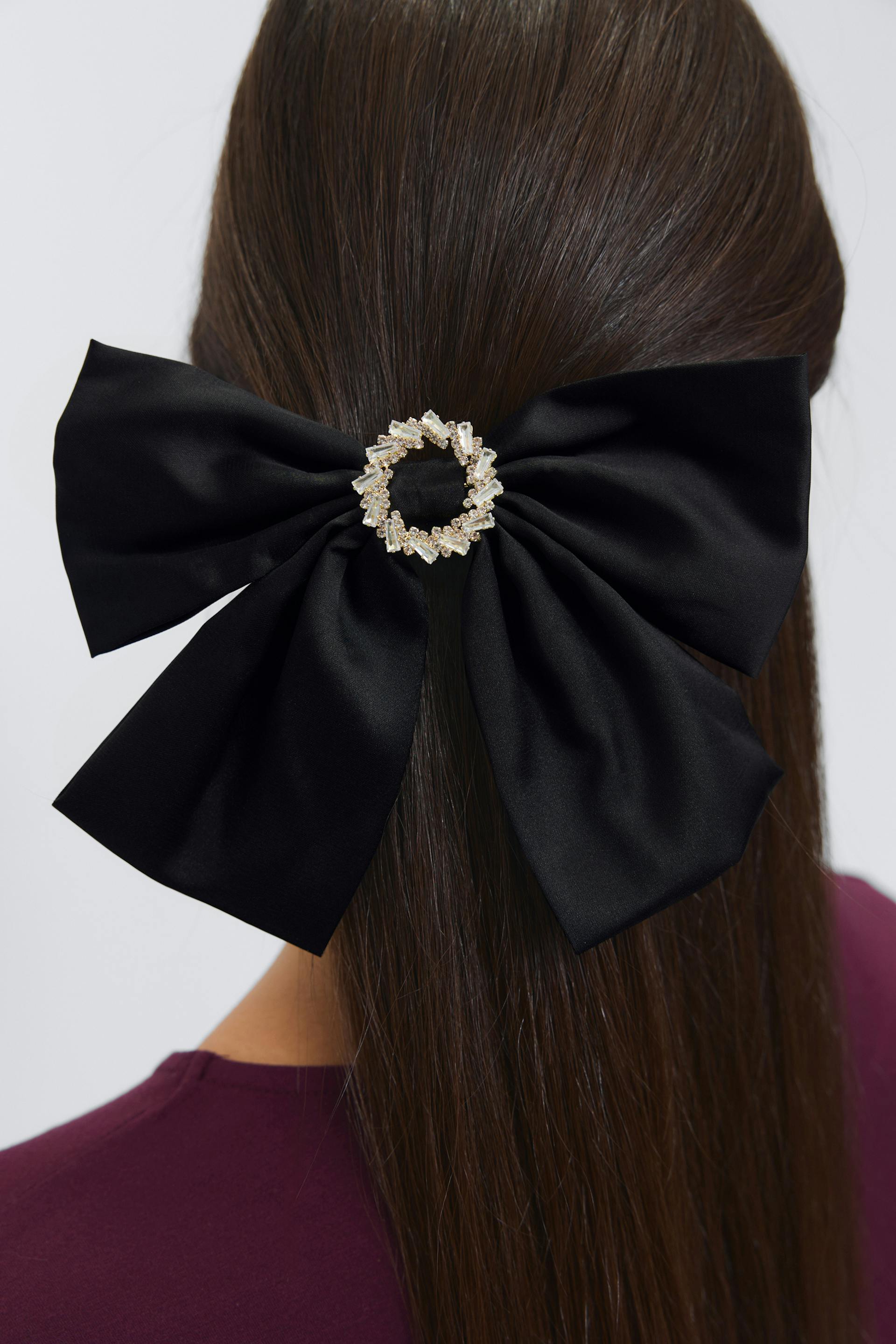 Black satin satin bow hair accessory with a circular crystal‑embellished gold brooch at the center, paired with a burgundy crew‑neck top.