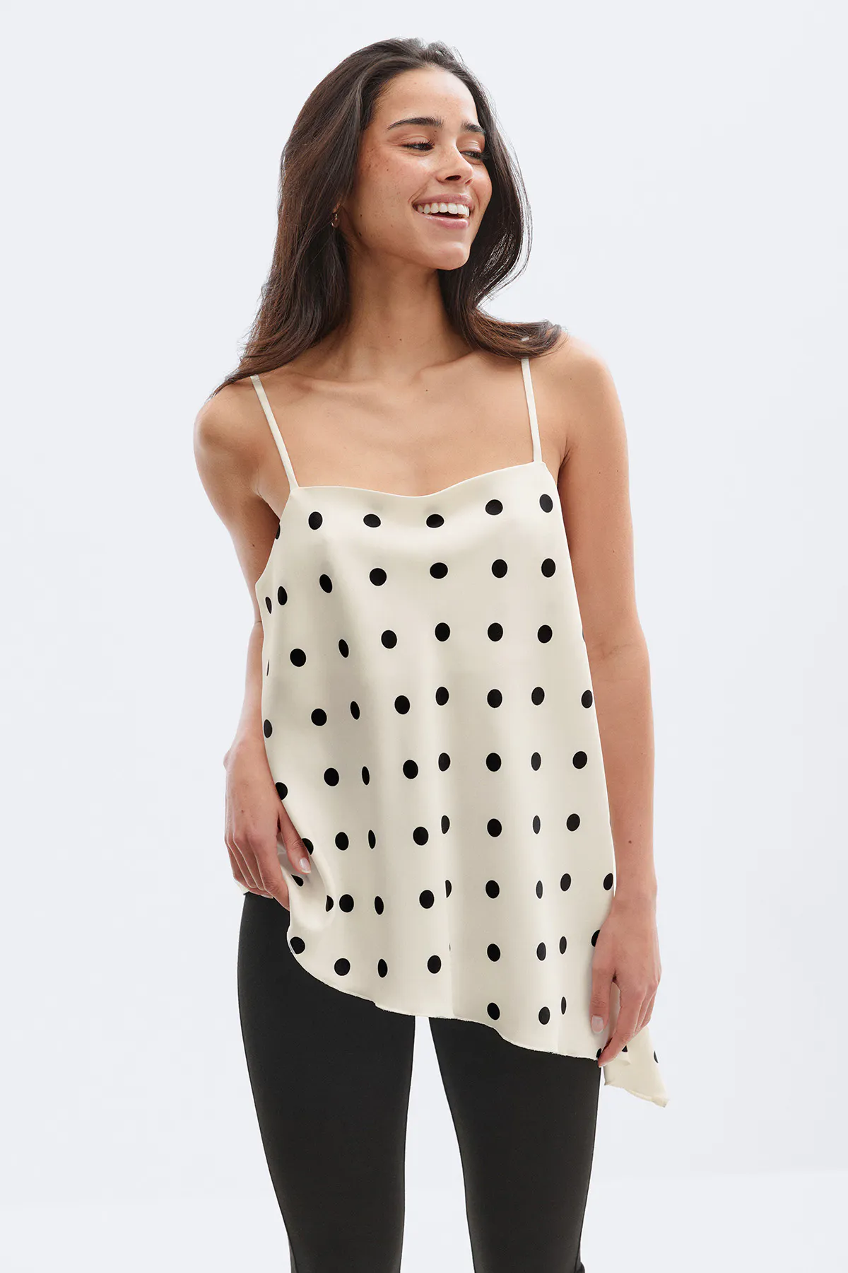 Norsunluunvärinen satiininen camisole, jossa on mustia pilkkuja, ohuet spagettihihnat ja epäsymmetrinen helma, yhdistettynä mustiin legginsseihin.