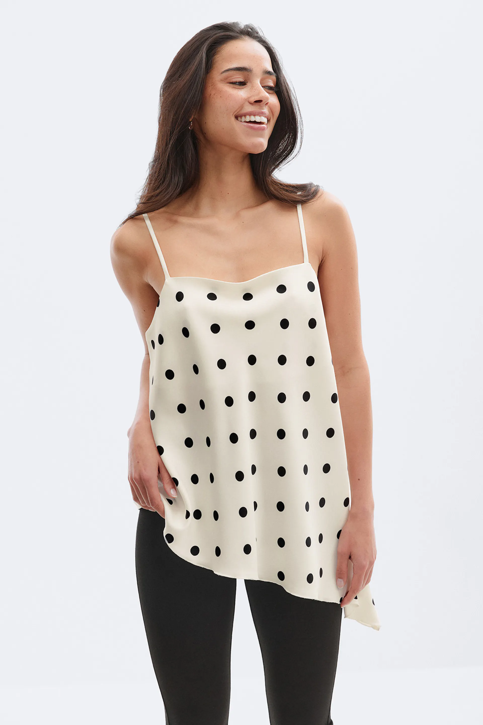 Norsunluunvärinen satiininen camisole, jossa on mustia pilkkuja, ohuet spagettihihnat ja epäsymmetrinen helma, yhdistettynä mustiin legginsseihin.