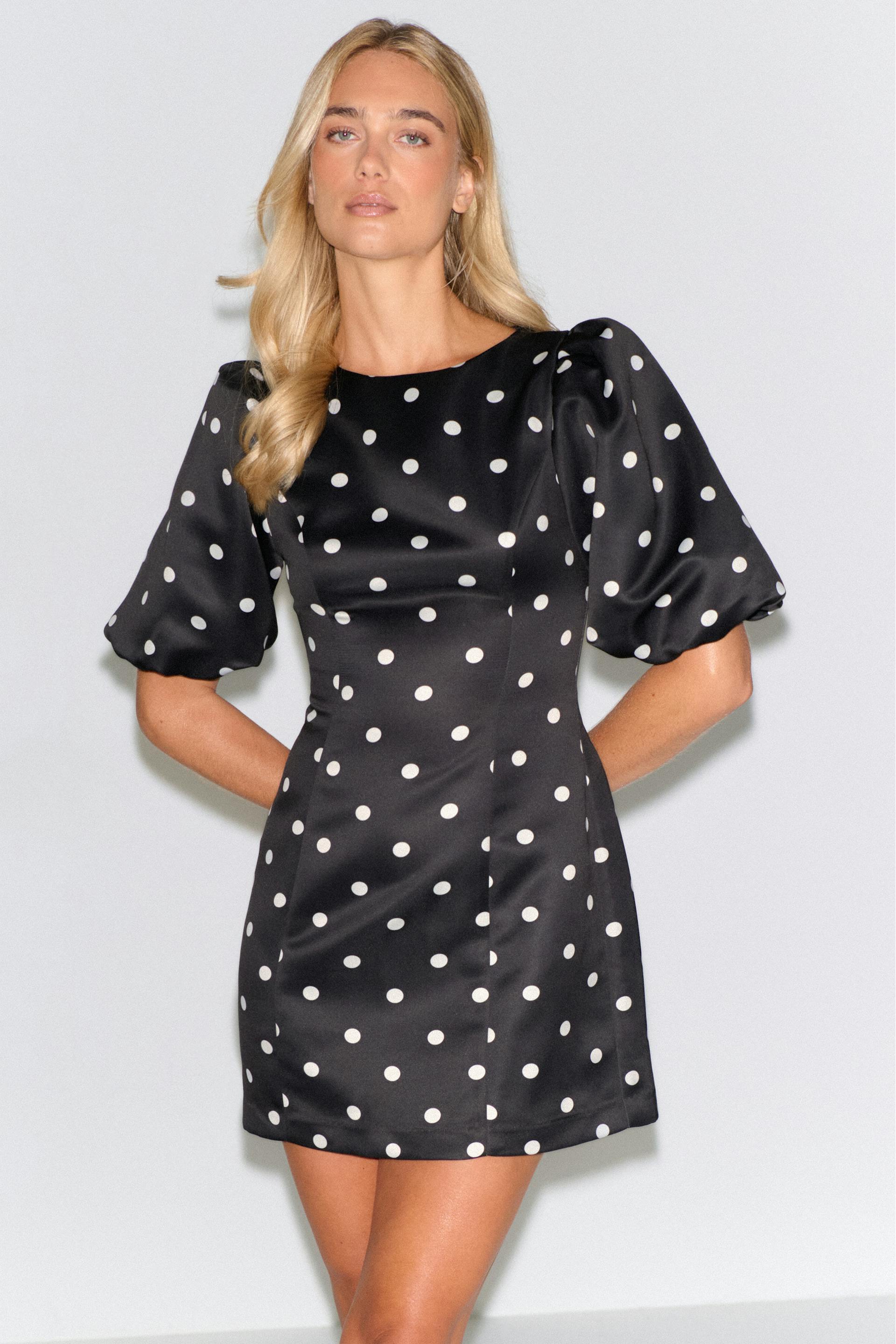 Miniklänning i svart satin med vita prickar, rund halsringning, figursydd överdel, A-linjeformad kjol och korta puffärmar med armbågslängd.