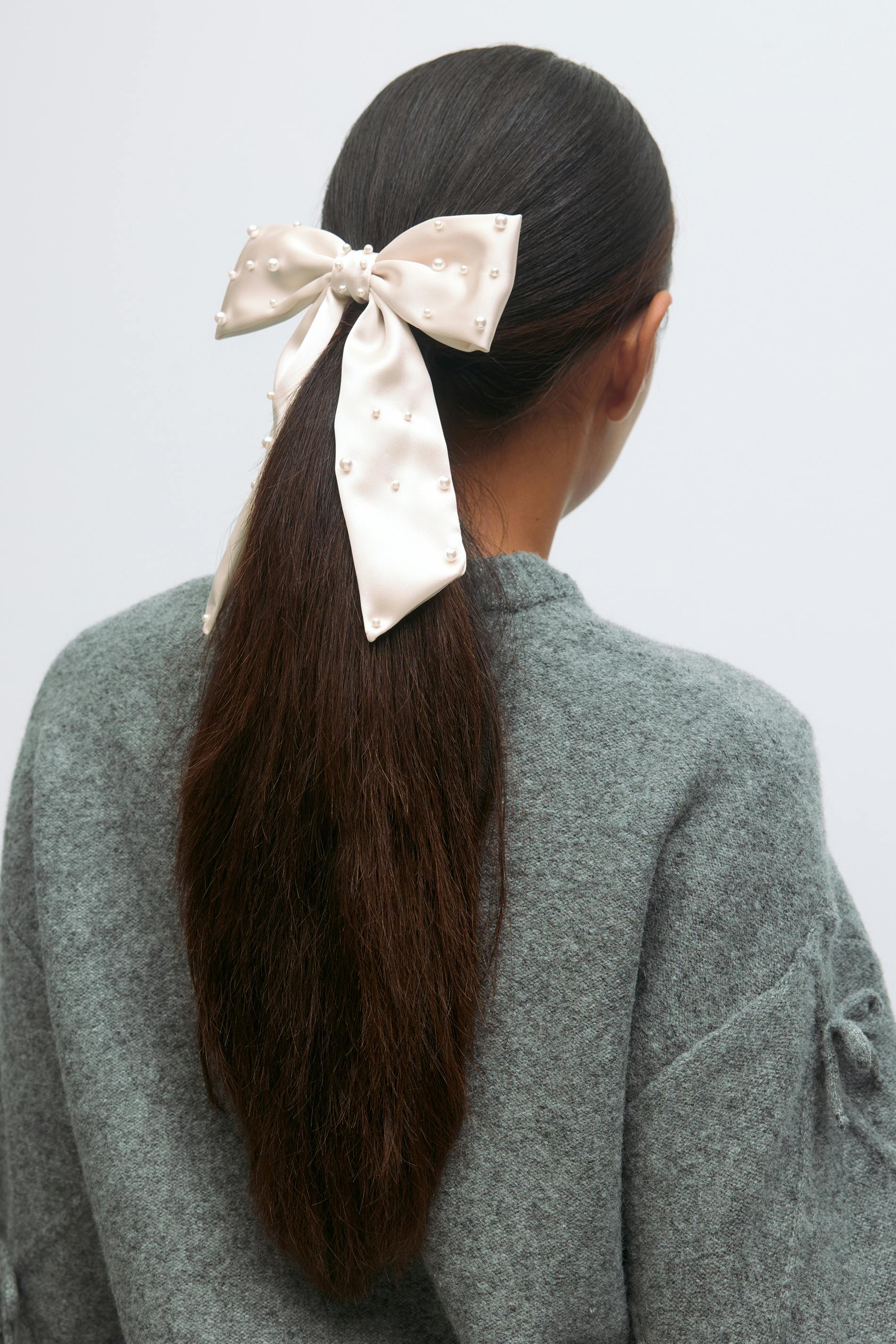 Ivory satin hair bow with scattered pearl embellishments, tied at the back of the head, paired with a grey textured knit sweater.