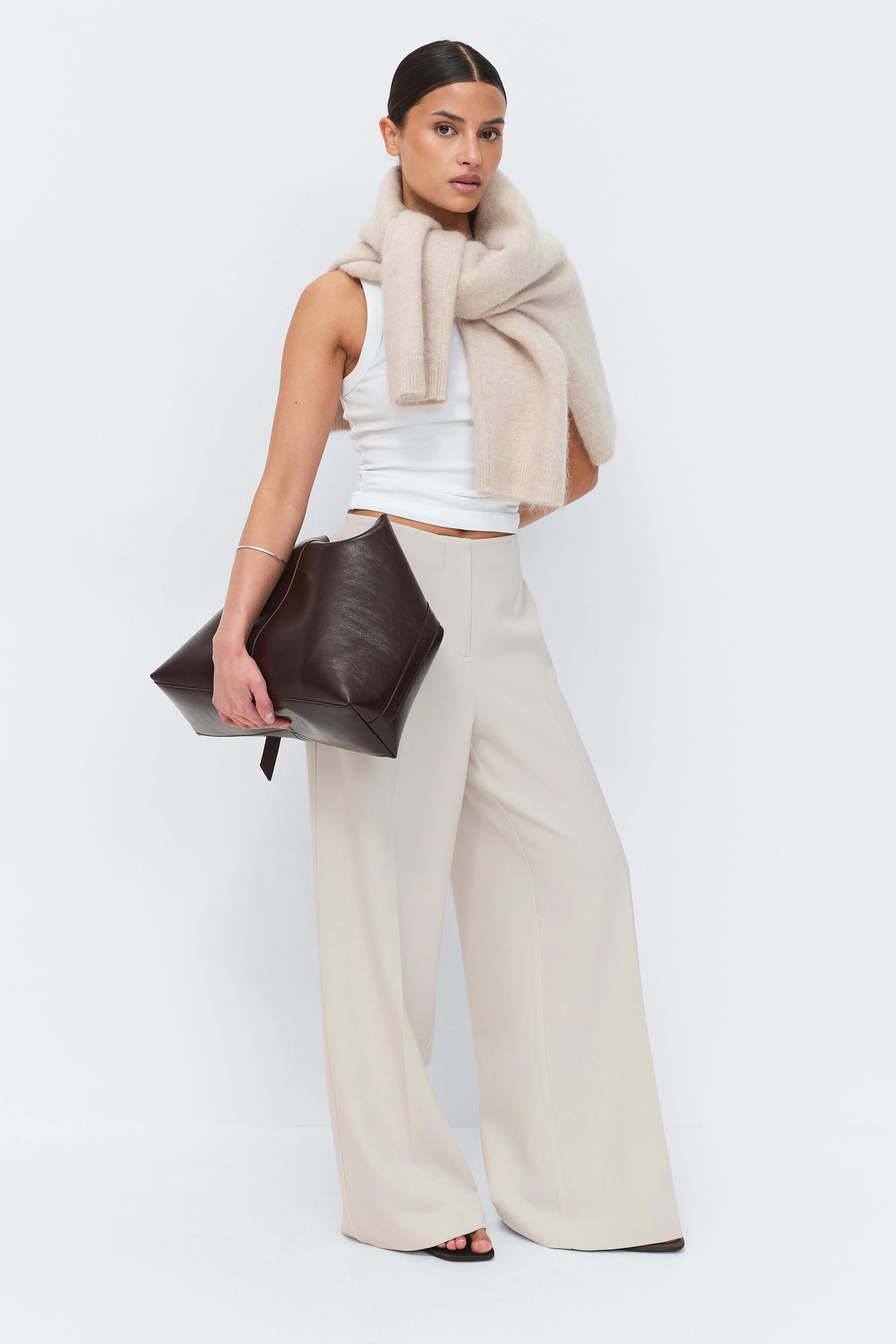 White fitted sleeveless tank top; high-waisted, wide-leg beige trousers; oversized beige knit scarf draped over the shoulders; dark brown leather oversized clutch; black open-toe sandals; slim silver bangle.