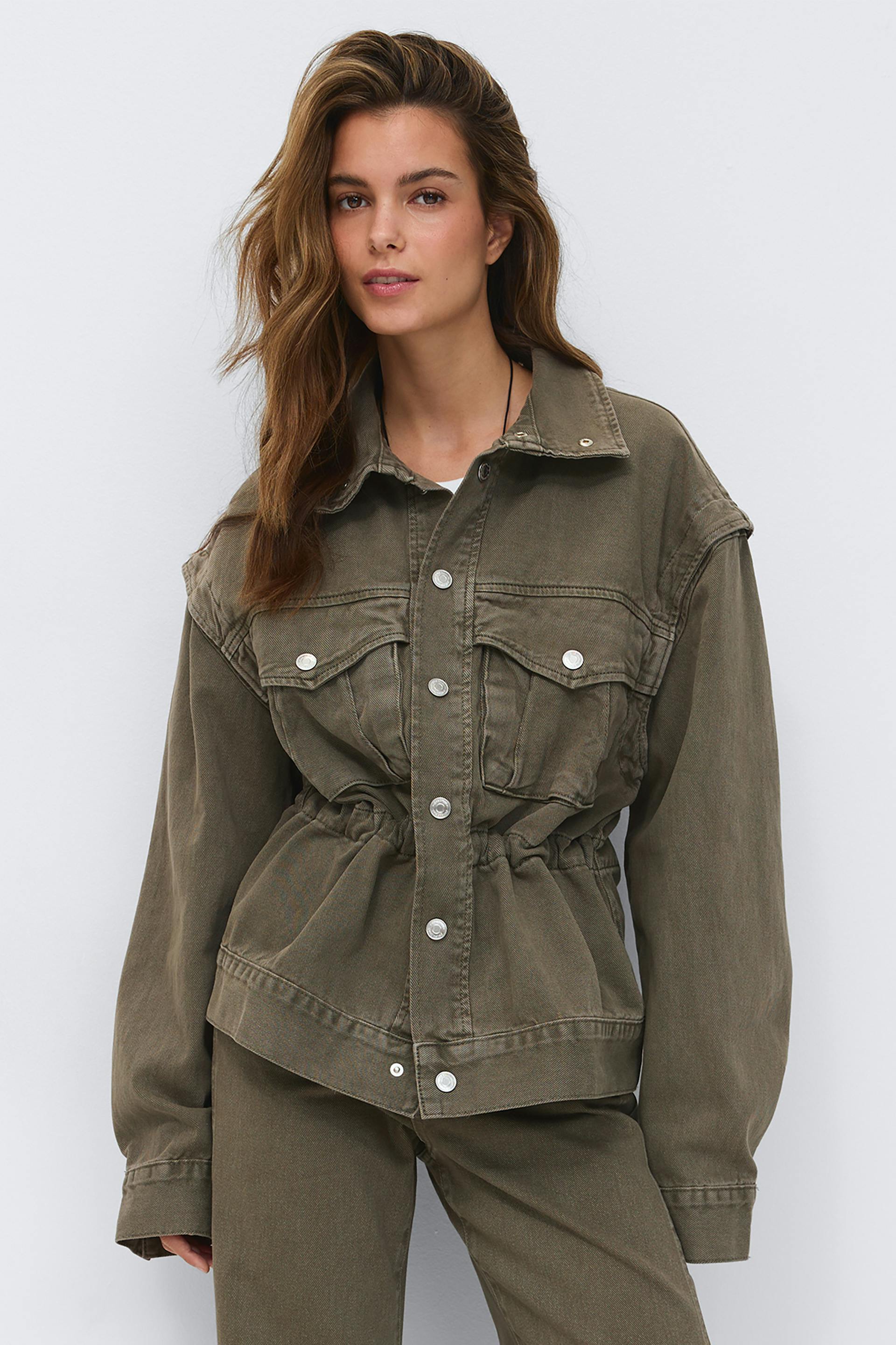 Oliivinvihreä pesty farkkutakki, jossa on hopeanväriset napit, läpälliset rintataskut ja vyötärönauha, yhdistettynä yhteensopiviin oliivinvihreisiin farkkuhousuihin.