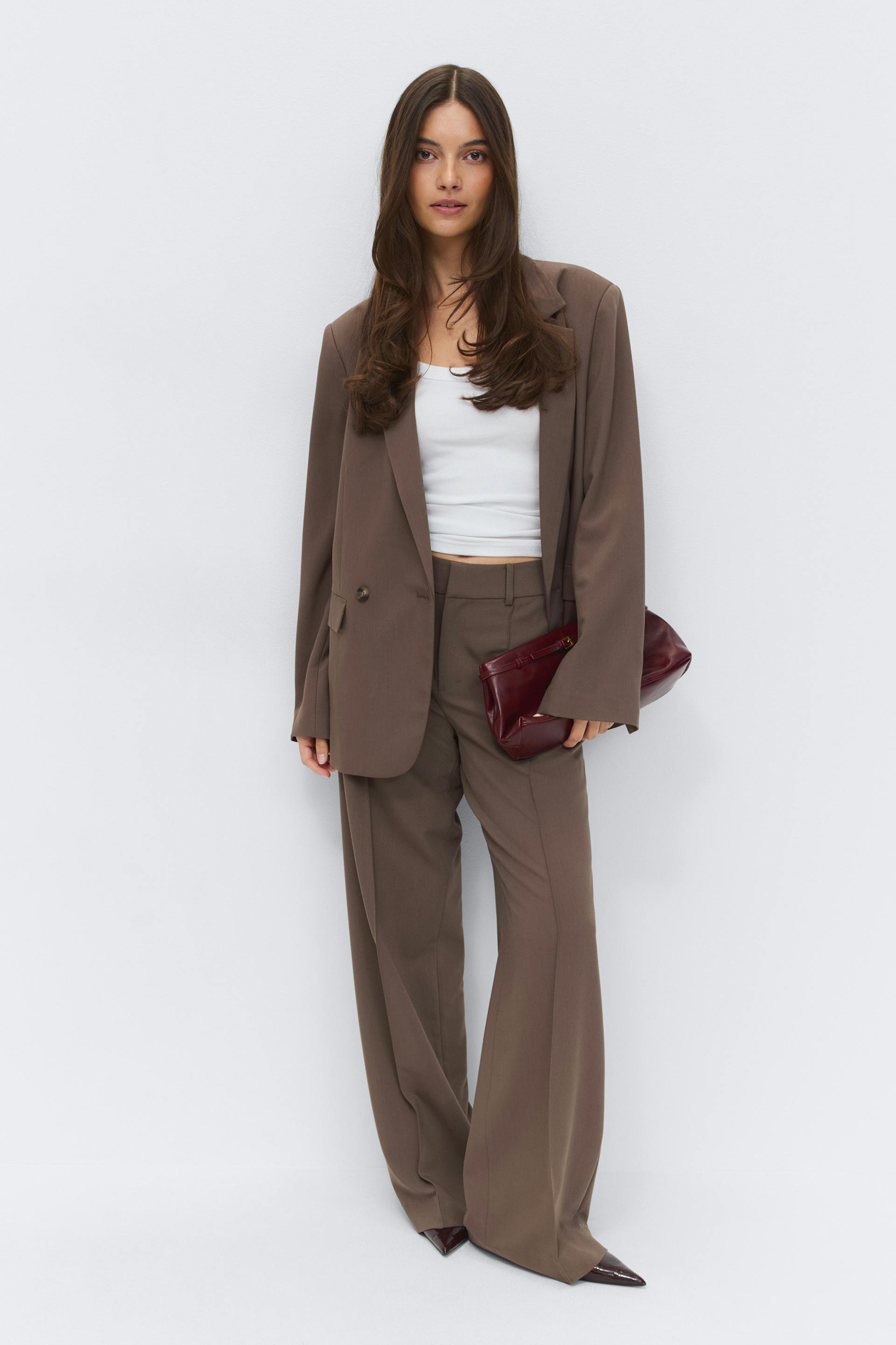 Model wearing a taupe-brown oversized single-breasted blazer with matching high-waisted wide-leg trousers, a white cropped tank top, a burgundy patent clutch and burgundy pointed-toe shoes.