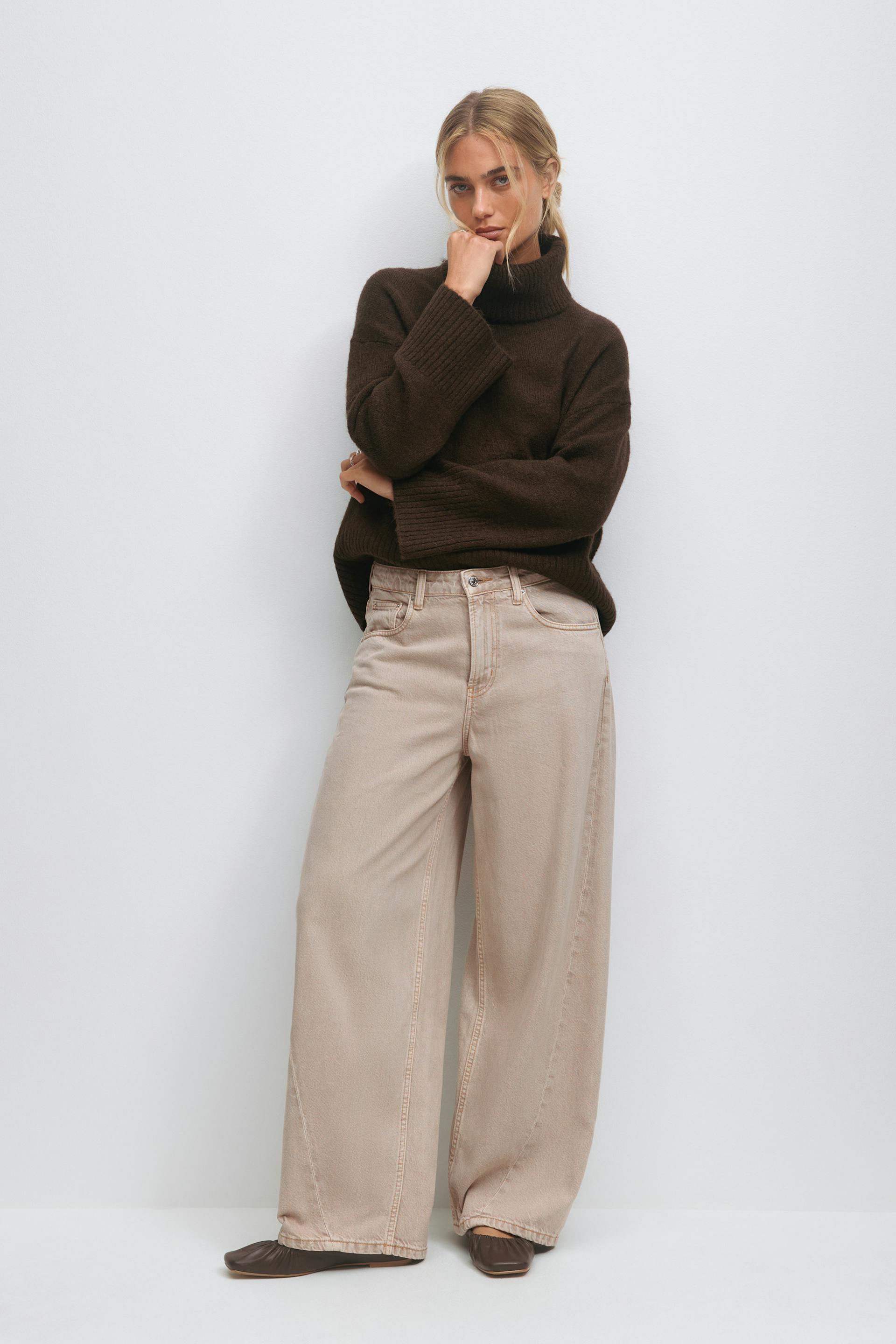 Dunkelbrauner, übergroßer Rollkragenpullover mit gerippten Bündchen, kombiniert mit beigefarbenen, weit geschnittenen Jeans mit hoher Taille und braunen, flachen Schuhen.