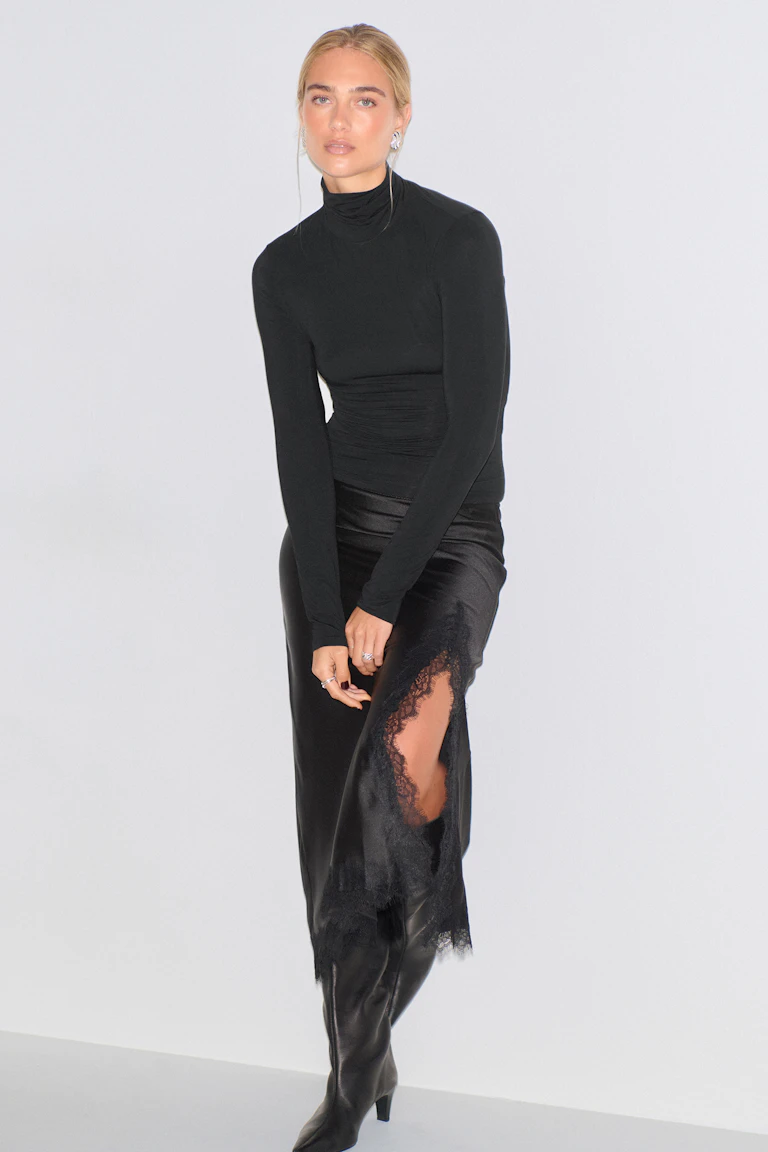 Fitted black long-sleeve turtleneck top paired with a black satin midi skirt featuring lace trim and a high side slit, styled with black knee-high leather boots.