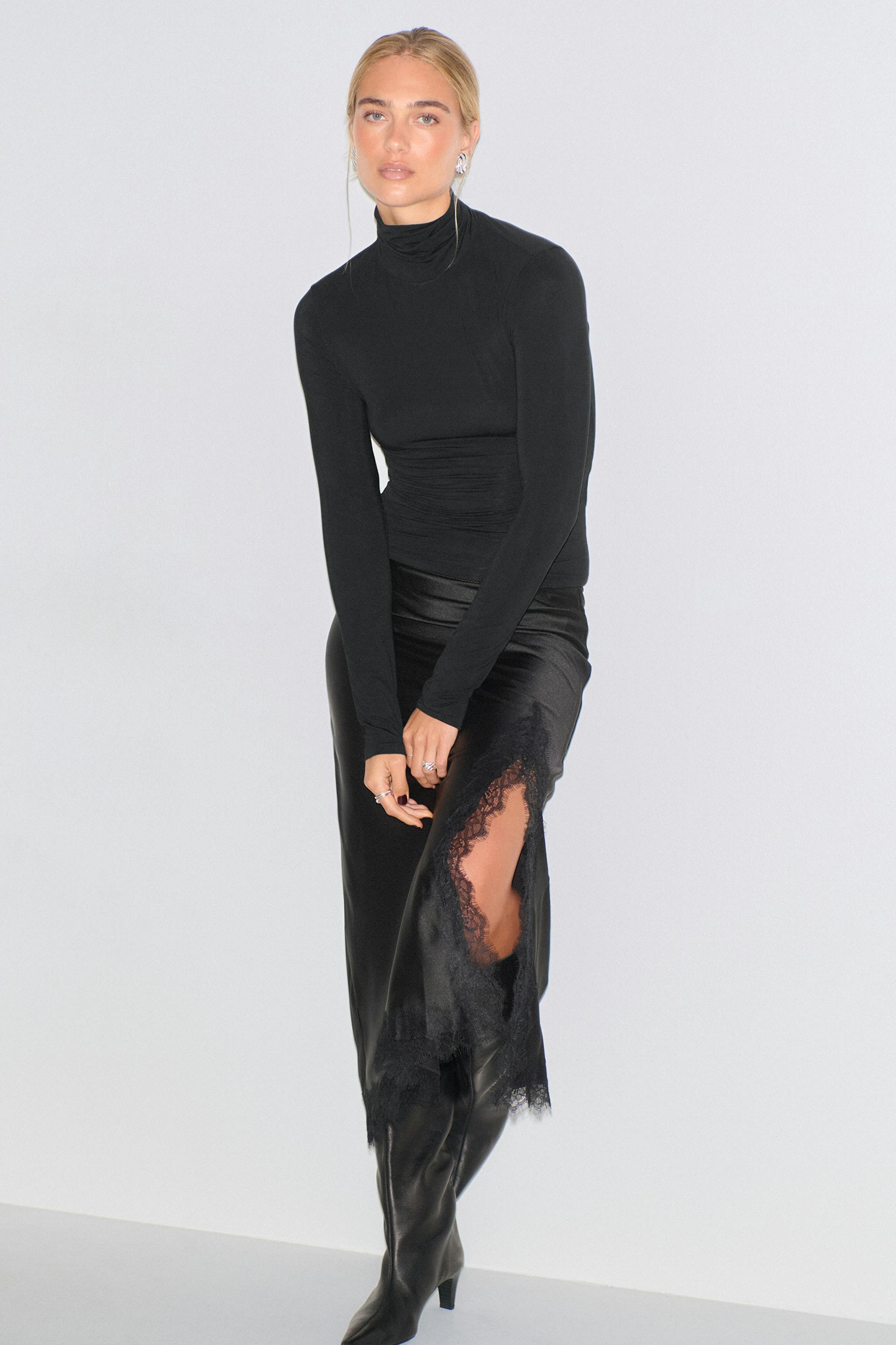 Schwarzes, tailliertes, langärmeliges Rollkragenoberteil, kombiniert mit einem schwarzen Midirock aus Satin mit Spitzenbesatz und hohem Seitenschlitz, dazu schwarze, kniehohe Lederstiefel.