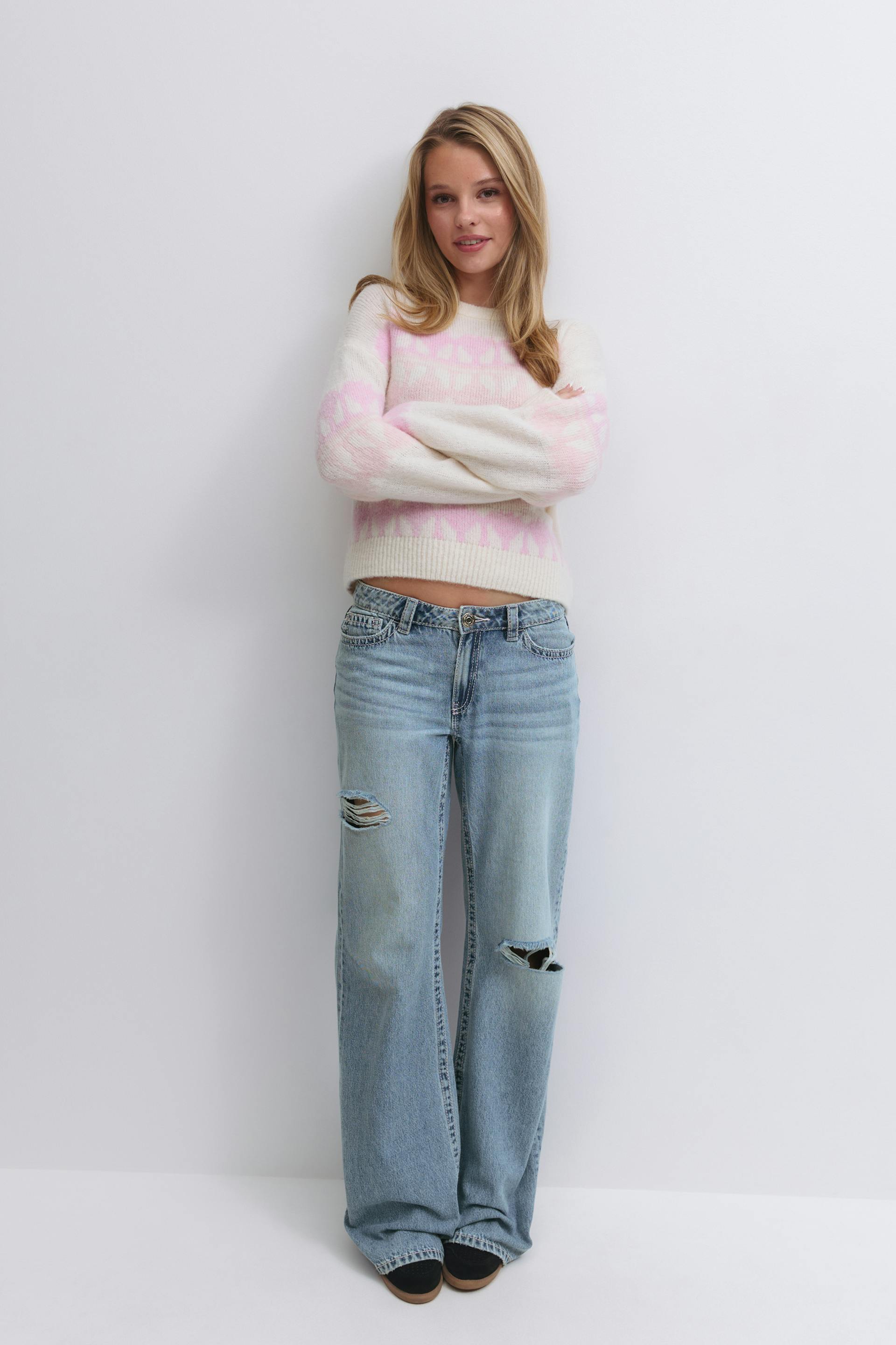 Woman wearing a cream and pale-pink patterned crewneck sweater, light-wash distressed wide-leg jeans with knee rips, and black slip-on shoes.