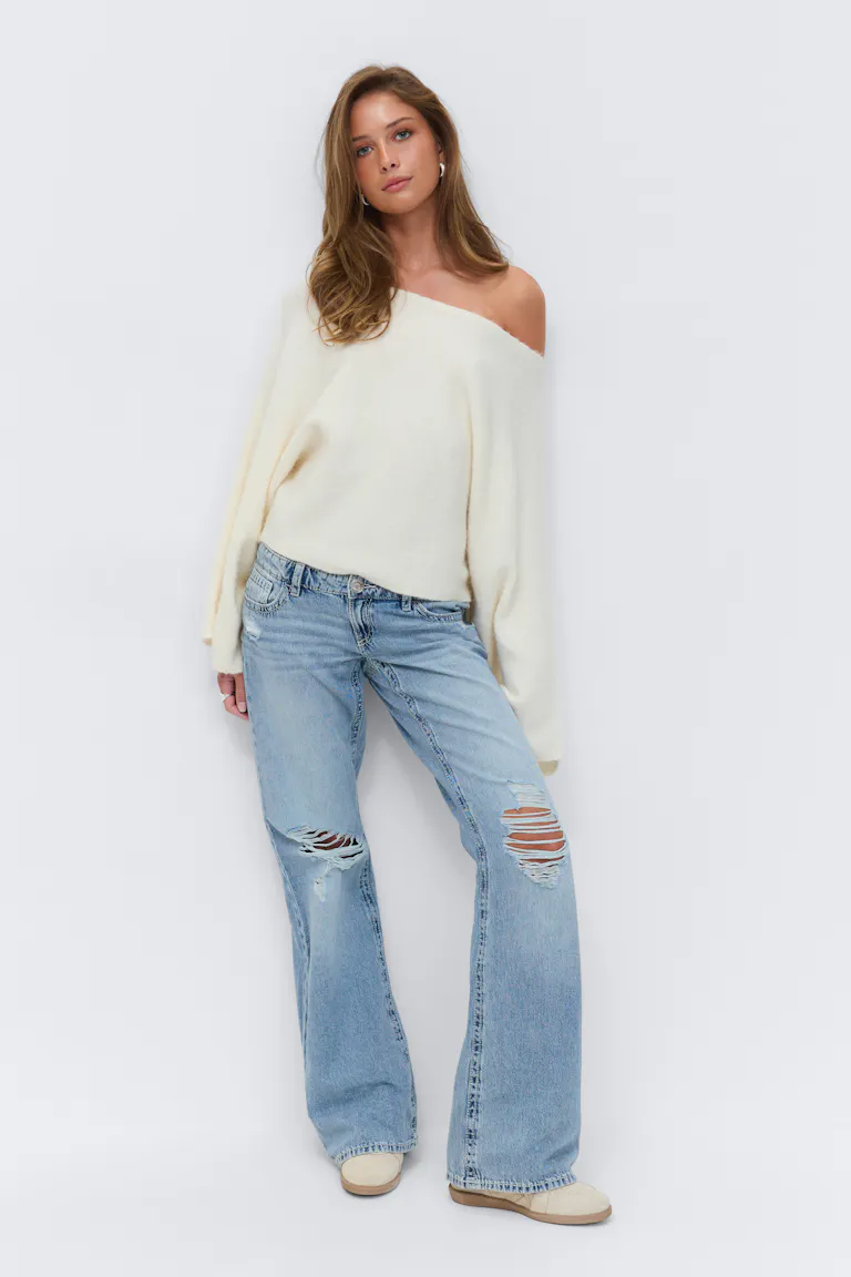 Pull à manches longues crème, jean large délavé avec déchirures au genou, baskets beiges et petites boucles d'oreilles.