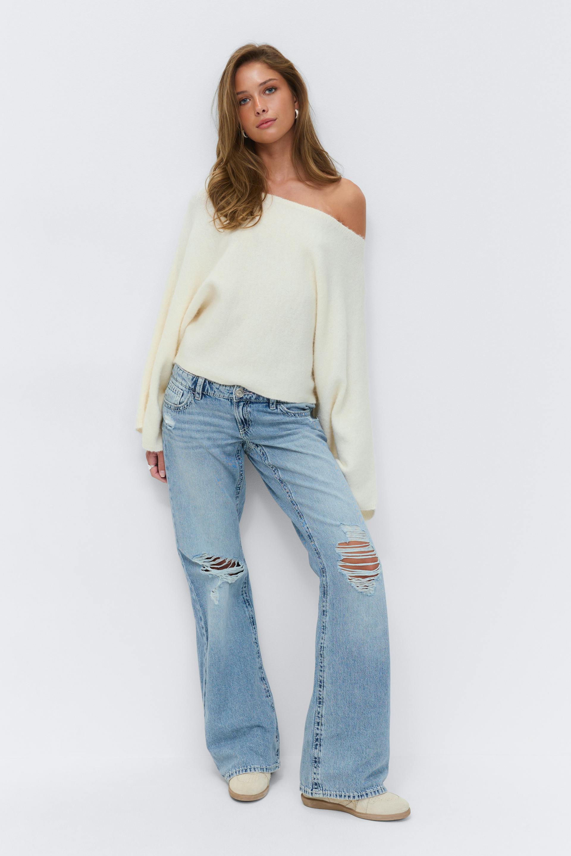 Kremfarget langermet genser uten skuldre, lyse, slitne jeans med vidde ben og slitasje på knærne, beige joggesko med lav topp og små øreringer.