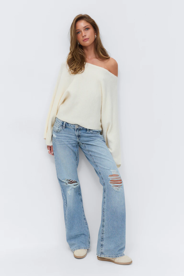 Cremefarbener, schulterfreier, langärmeliger Pullover, hellgewaschene Jeans mit weitem Bein und Knierissen, beigefarbene Low-Top-Sneakers und kleine Ohrringe.