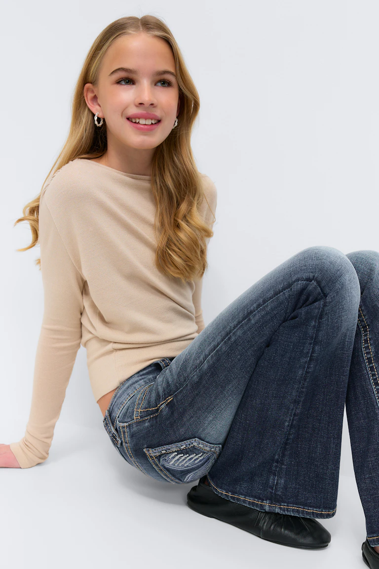 Beige trui met lange mouwen, donker gewassen flared jeans met geborduurde achterzakken, zwarte ballerina's en zilveren oorbellen.