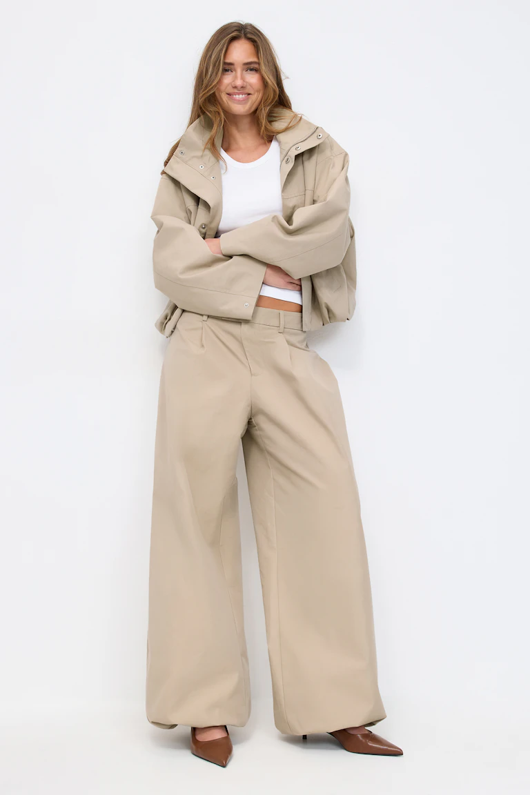 Beige übergroße Kurzjacke mit Druckknöpfen, weißes Tank mit Rundhalsausschnitt, beige Hose mit weitem Bein und braune spitze Absätze.