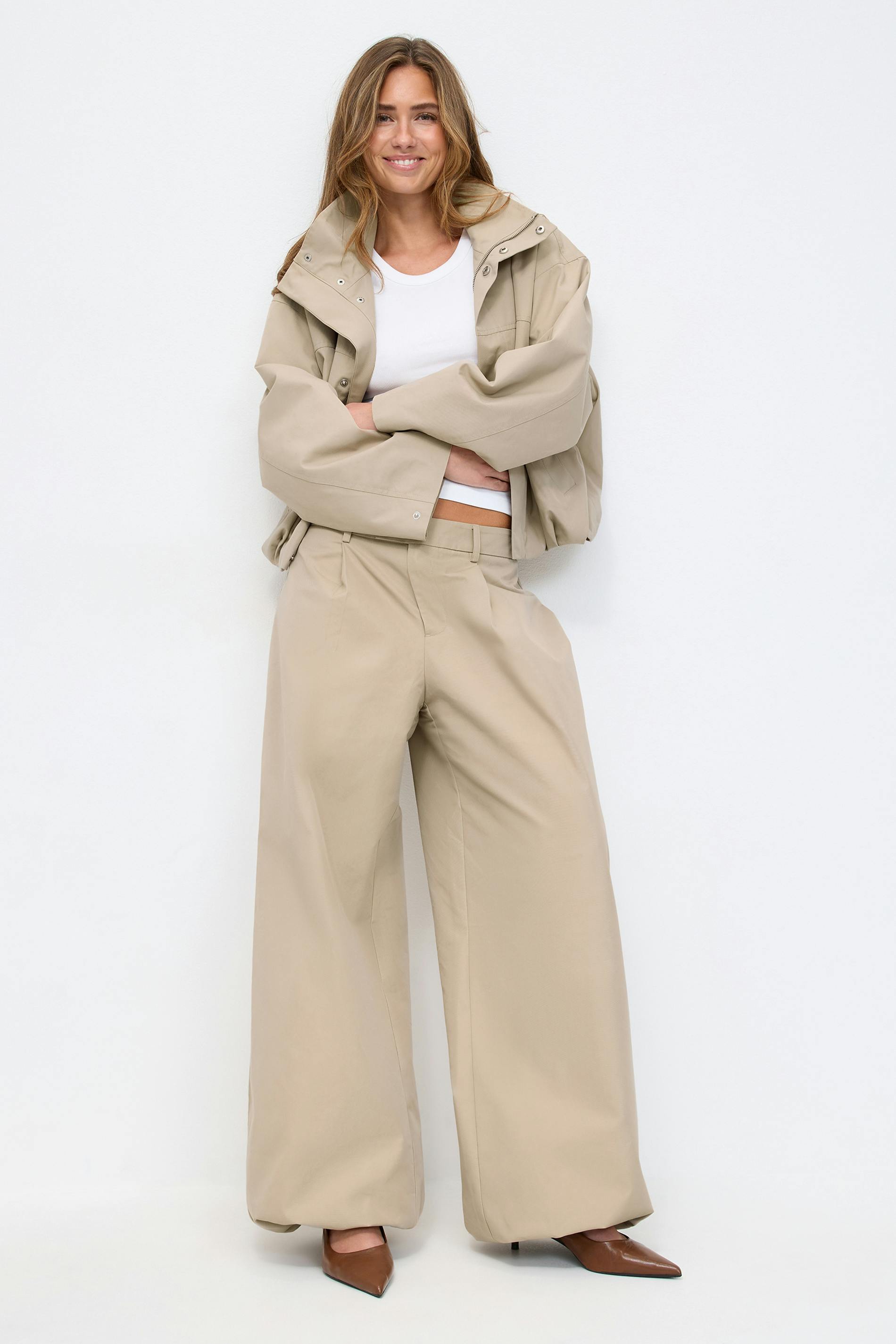 Oversized beige croppad jacka med tryckknappar, vitt rundhalsat linne, beige byxor med vida ben och hög midja samt bruna spetsiga klackar.