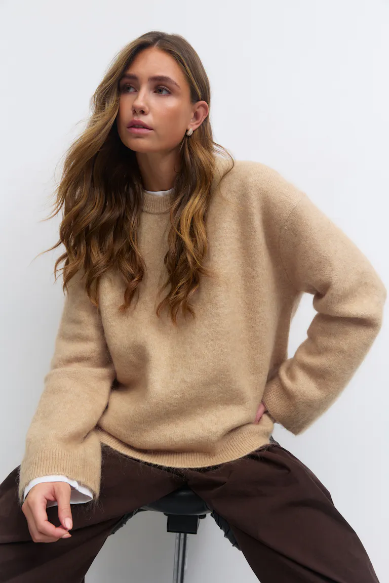 Beigefarbener, übergroßer, flauschiger Pullover mit Rundhalsausschnitt über einem weißen T-Shirt mit Rundhalsausschnitt; dunkelbraune, locker sitzende Hose; kleine goldene Ohrringe.