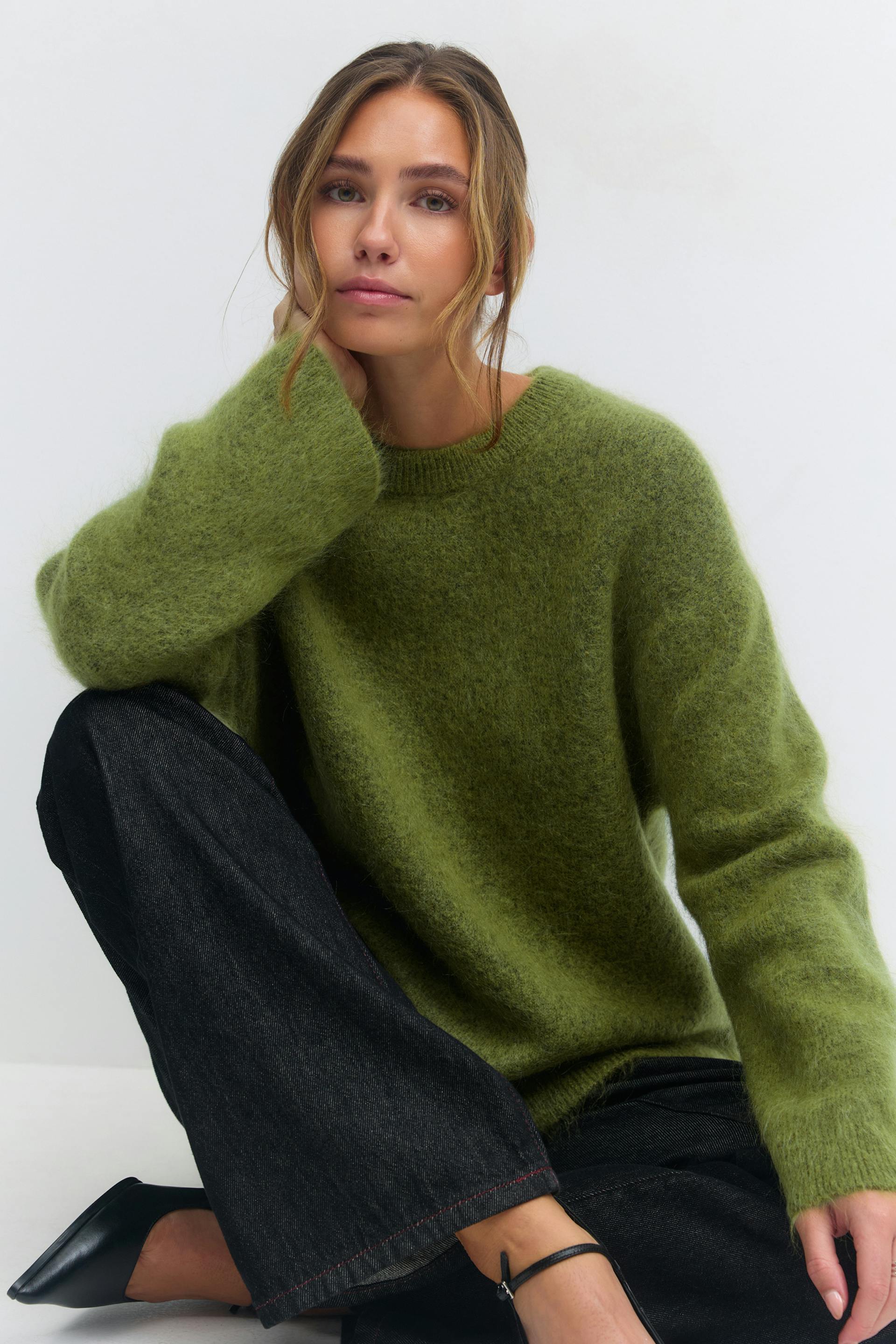 Olivgrüner, flauschiger Pullover mit Rundhalsausschnitt, schwarze Jeans mit weitem Bein und schwarze, spitze Riemchen-Absätze.