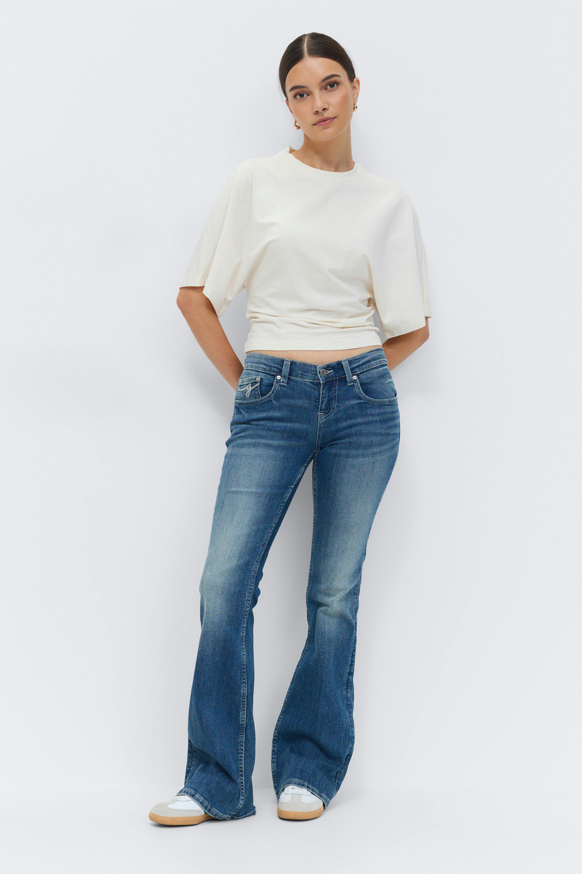 Ivory short-sleeve cropped top with gathered waist; medium-wash flared jeans; white sneakers with gum soles; small gold hoop earrings.