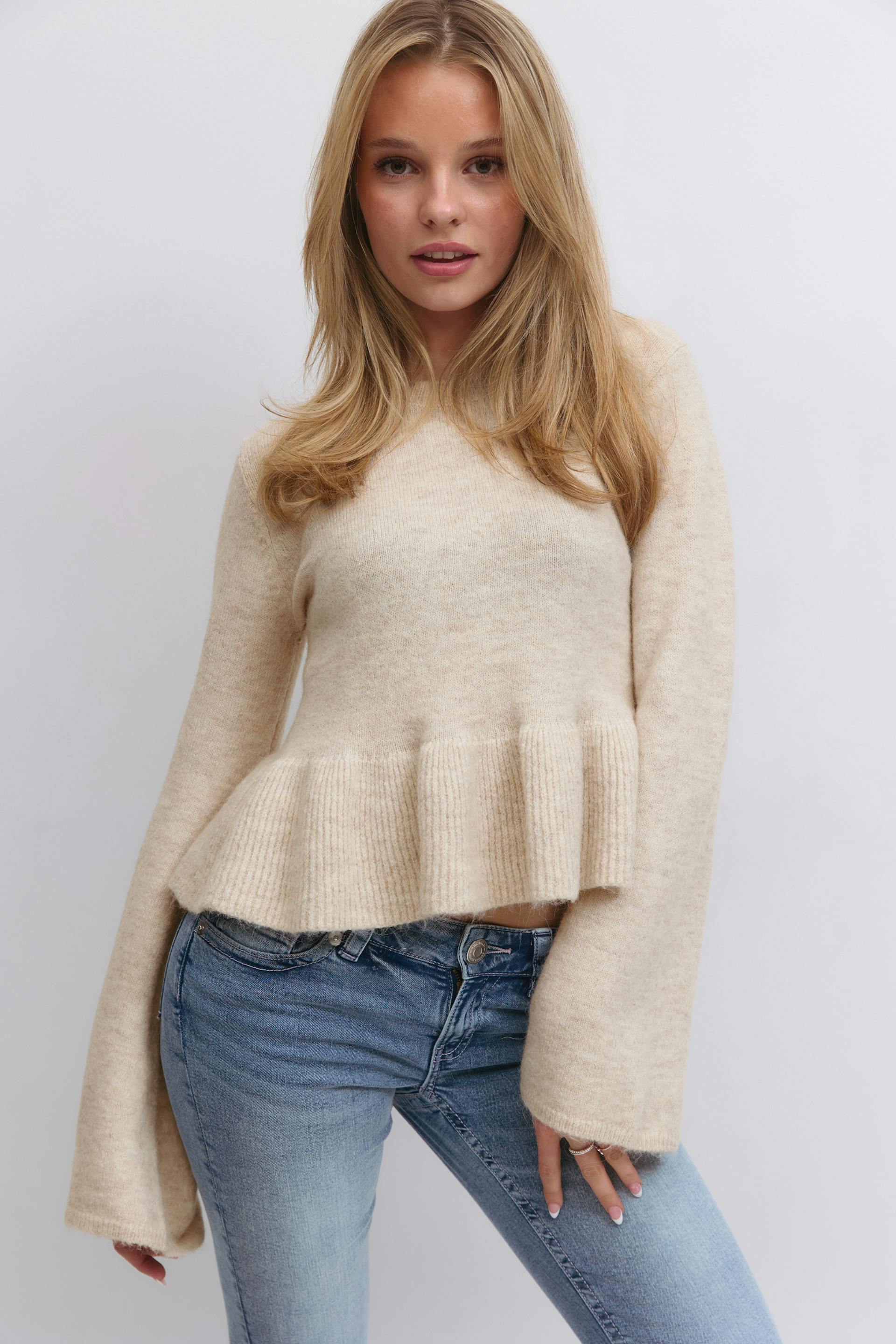 Cream ribbed peplum knit sweater with long bell sleeves, paired with light‑wash skinny jeans.