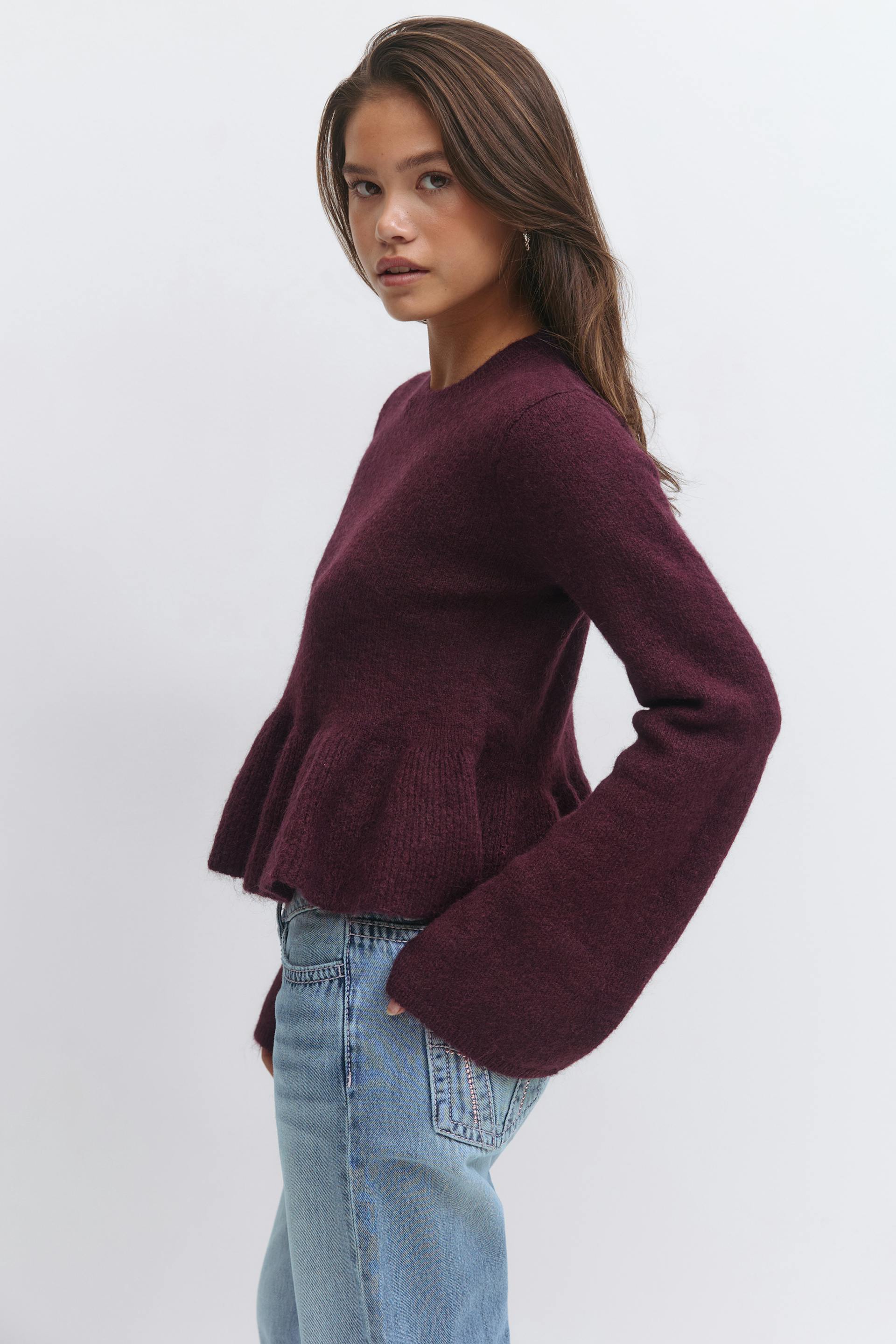 Deep burgundy textured knit sweater with a fitted bodice, peplum hem and long bell sleeves, paired with light‑wash straight‑leg jeans.