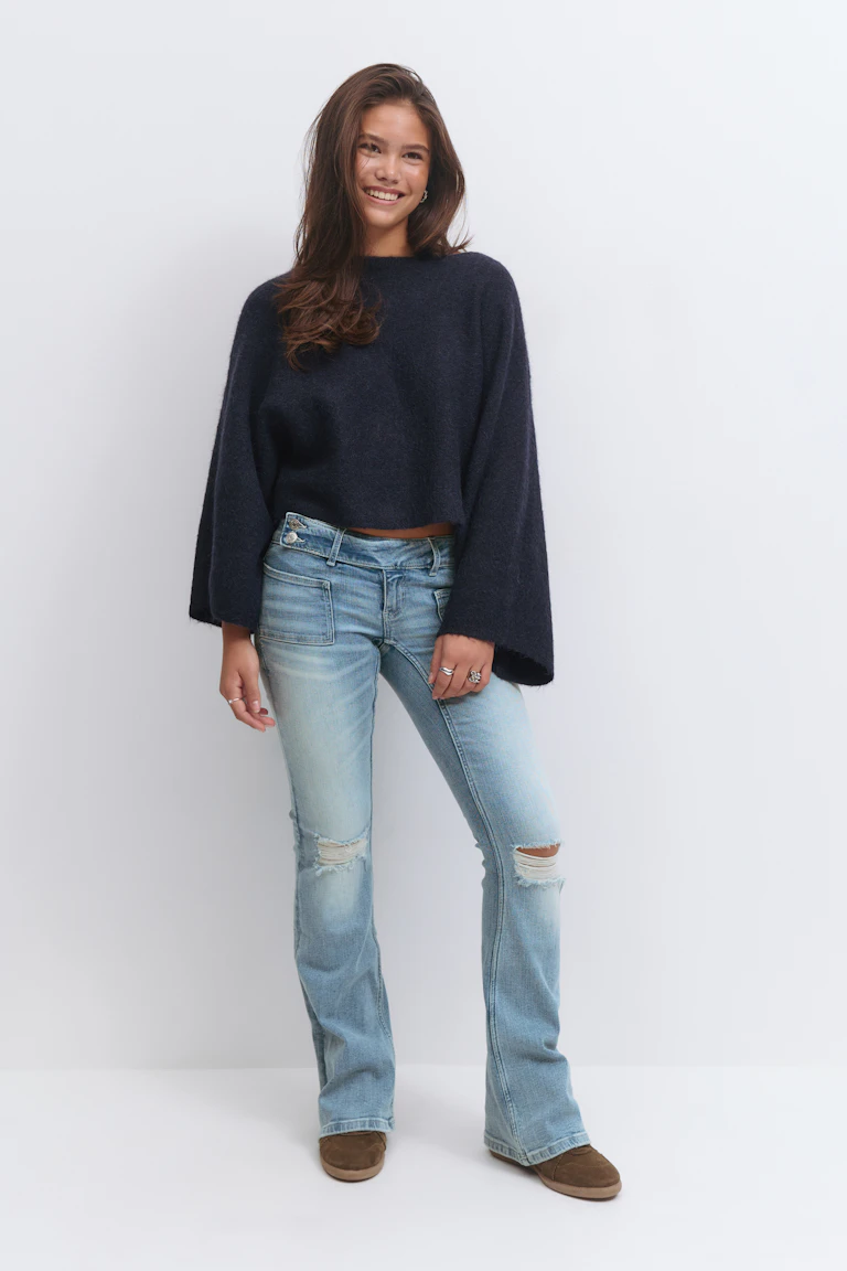 Marineblauer Pullover mit Rundhalsausschnitt und weiten Ärmeln; hell gewaschene Jeans mit Knierissen und aufgesetzten Taschen vorne; braune Wildlederschuhe zum Hineinschlüpfen.