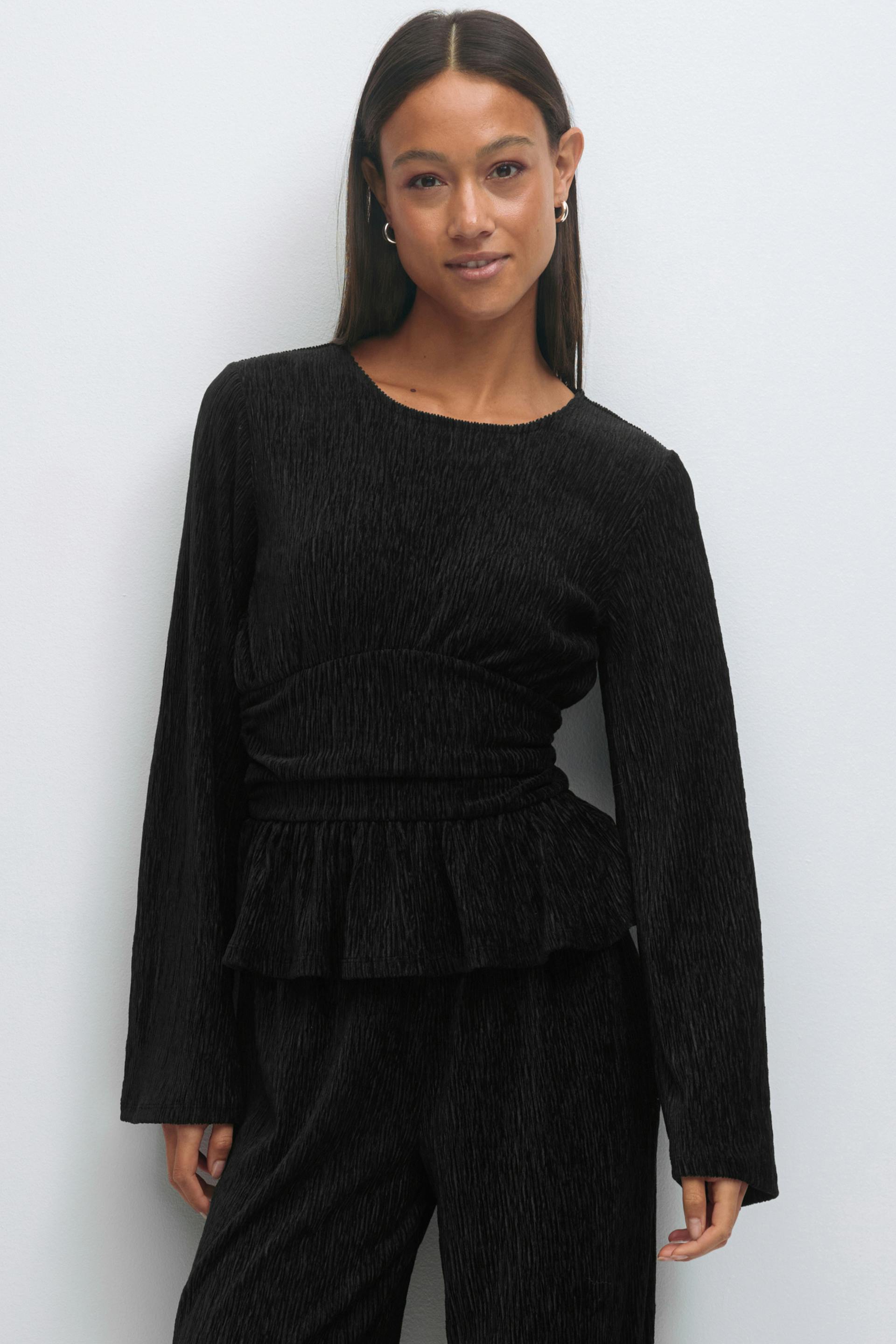 Black textured long-sleeve peplum top with round neckline and ruched, cinched waist, paired with matching wide-leg trousers; small silver hoop earrings.