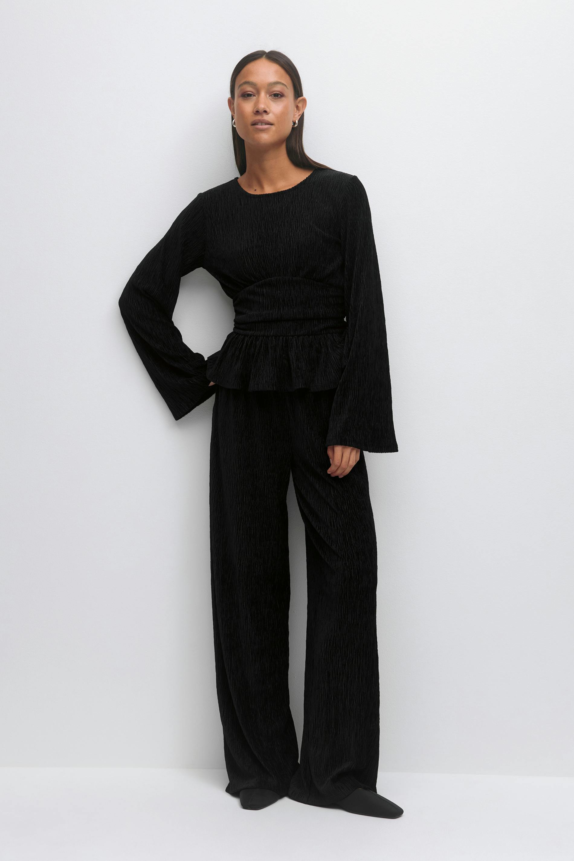 Woman wearing a black textured long-sleeve top with a gathered waist and peplum, paired with matching wide-leg trousers, black pointed flats and small hoop earrings.