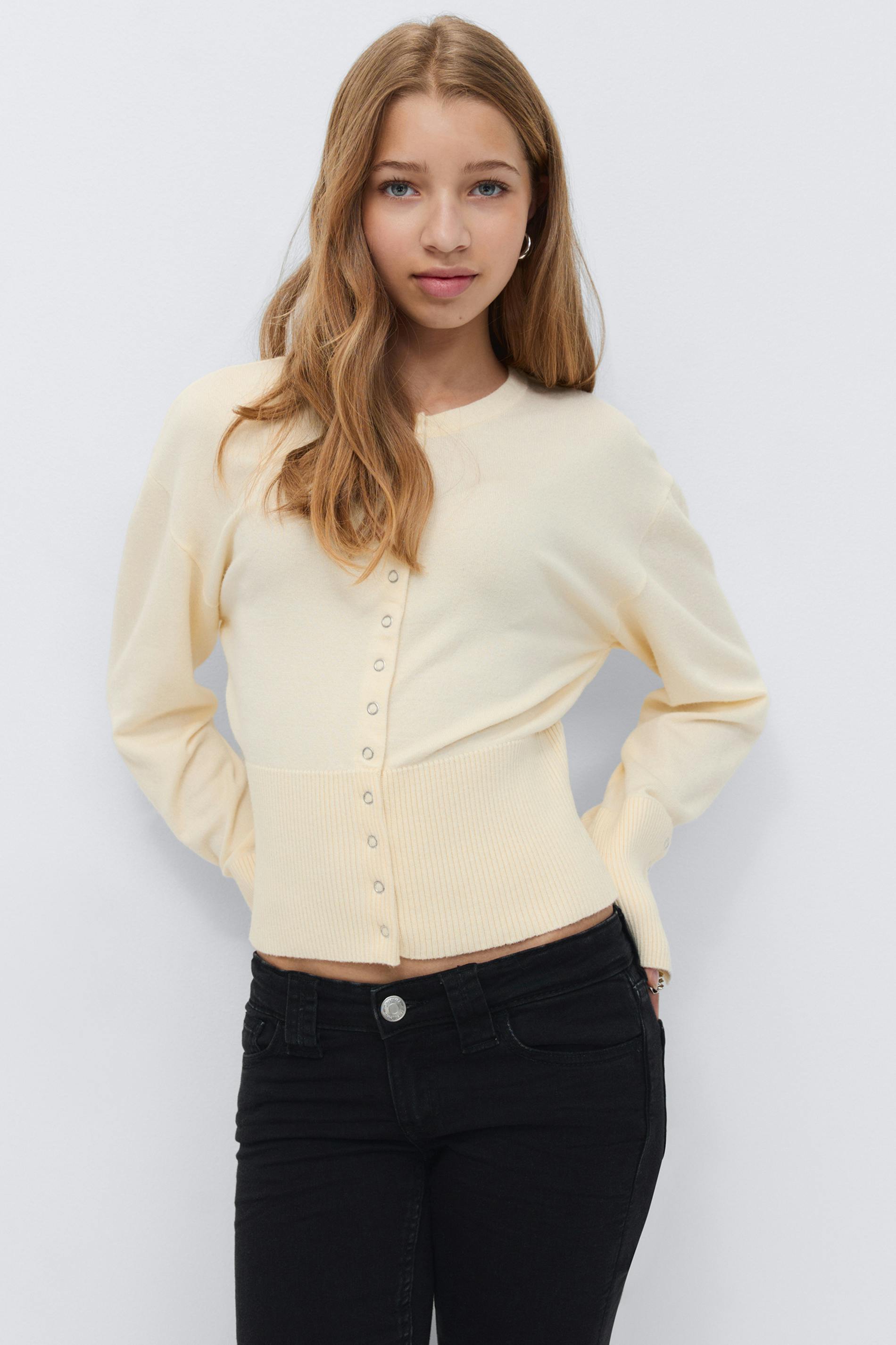 Cream cropped button-front cardigan with ribbed wide waistband and long sleeves, paired with black skinny jeans.