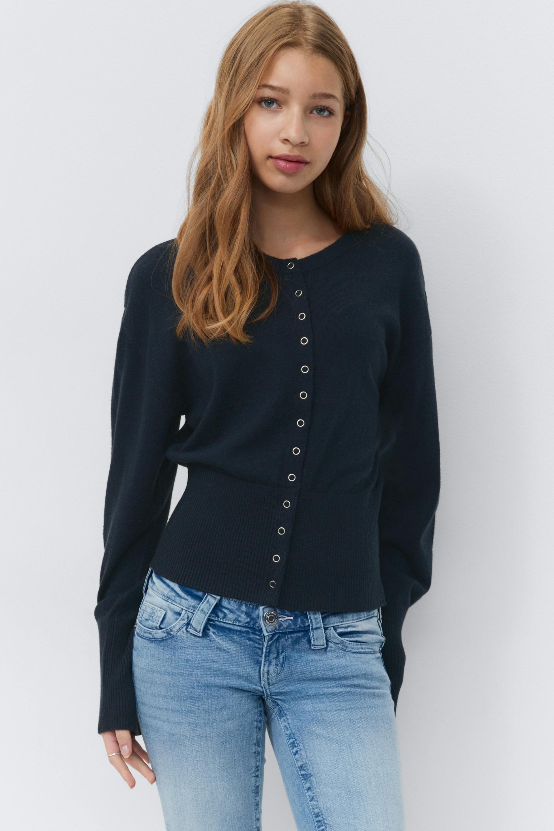 Navy long-sleeve button-front cardigan with metal grommet buttons and a ribbed, cinched waistband, paired with light-wash slim-fit jeans.