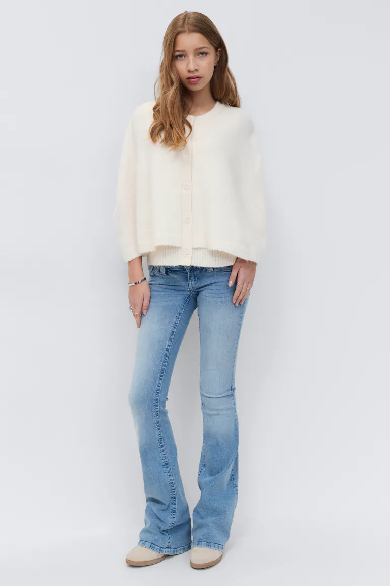 Cremefarbene Strickjacke mit Knopfleiste und Rippbündchen, hellen Schlagjeans, beigen Wildlederschuhen und silbernen Armbändern und Ringen.