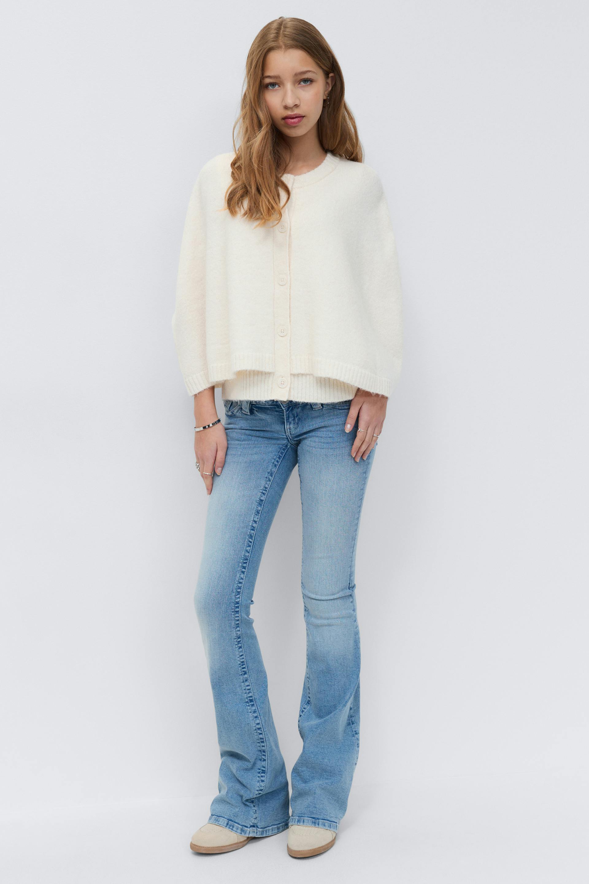 Cream button-front cropped cardigan with ribbed trim, light-wash flared jeans, beige suede slip-on shoes, and silver bracelets and rings.
