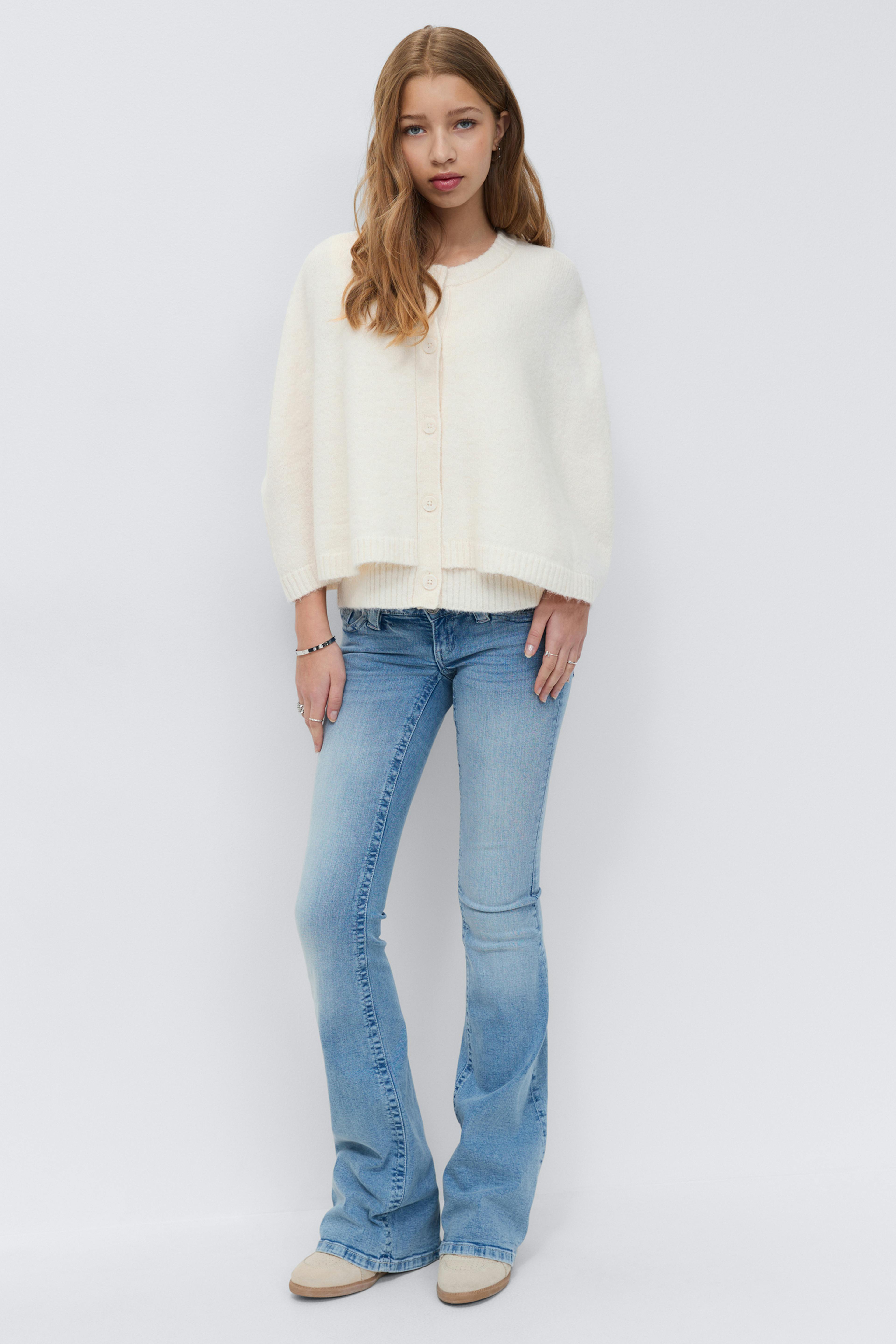 Crèmefarget kort cardigan med knapper foran og ribbestrikket kant, lyse jeans, beige semskede slip-on-sko og sølvfargede armbånd og ringer.