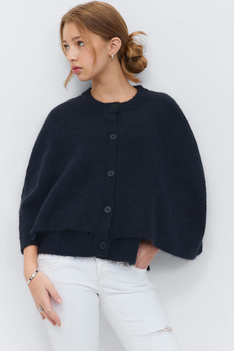 Het model draagt een donker marineblauw donzig capevest met knoopsluiting en geribde ronde hals, gecombineerd met witte distressed skinny jeans en zilveren oorbellen, ringen en een armband.